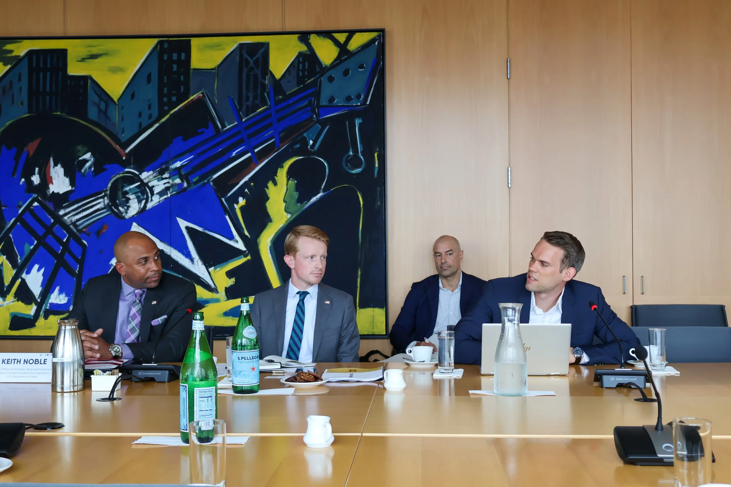 Four men in business suits sitting at a conference table in a meeting room, with a large abstract painting of a cityscape on the wall behind them and various beverages on the table.