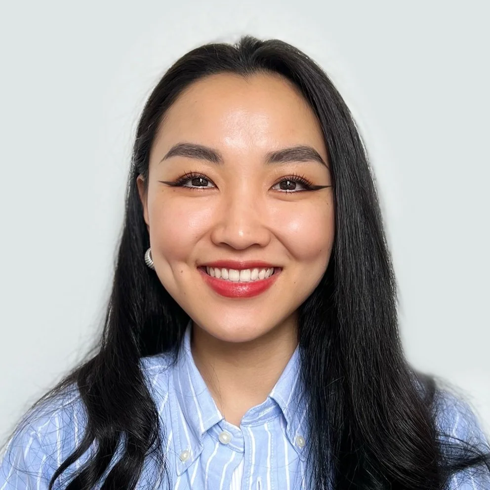 A young woman with long black hair, wearing makeup, and a blue and white striped button-up shirt, smiling at the camera against a plain white background.