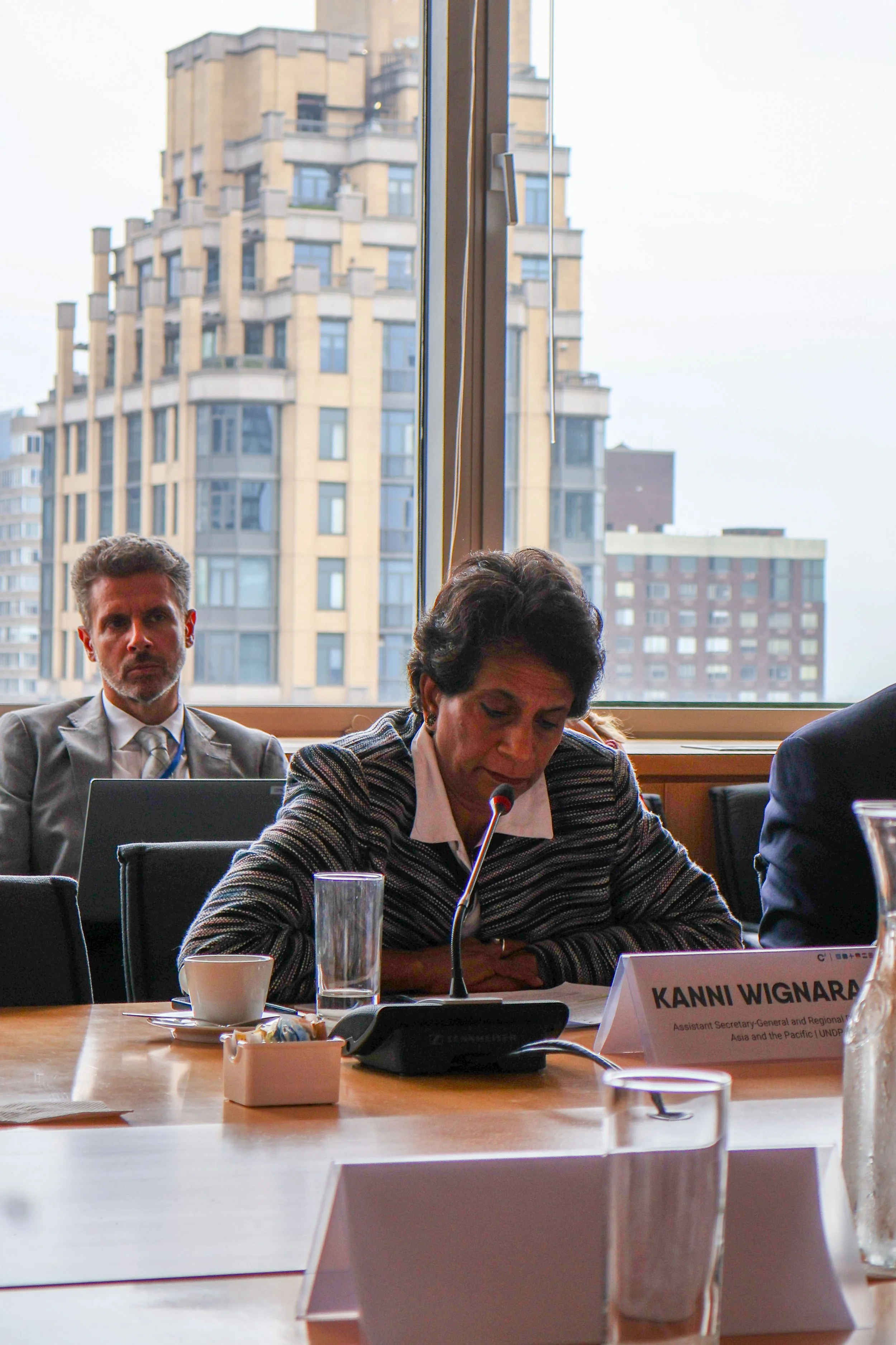 A woman is speaking into a microphone at a conference table, with a nameplate reading "Kanni Wignaraja" in front of her, and a man in a suit sitting behind her.