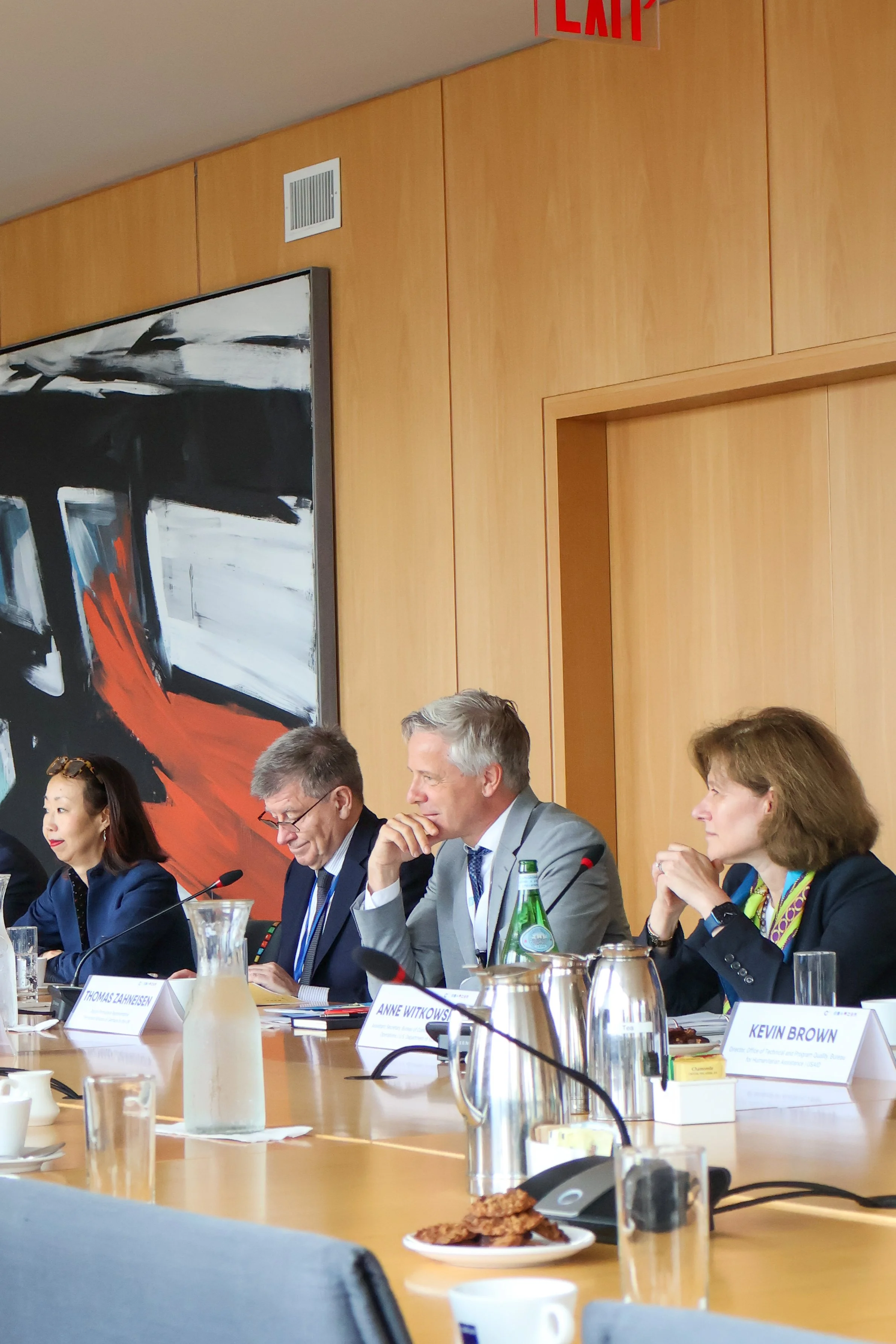 People sitting at a conference table with microphones, water bottles, and nameplates, engaged in a discussion in a modern conference room with wood paneling and abstract art on the wall.