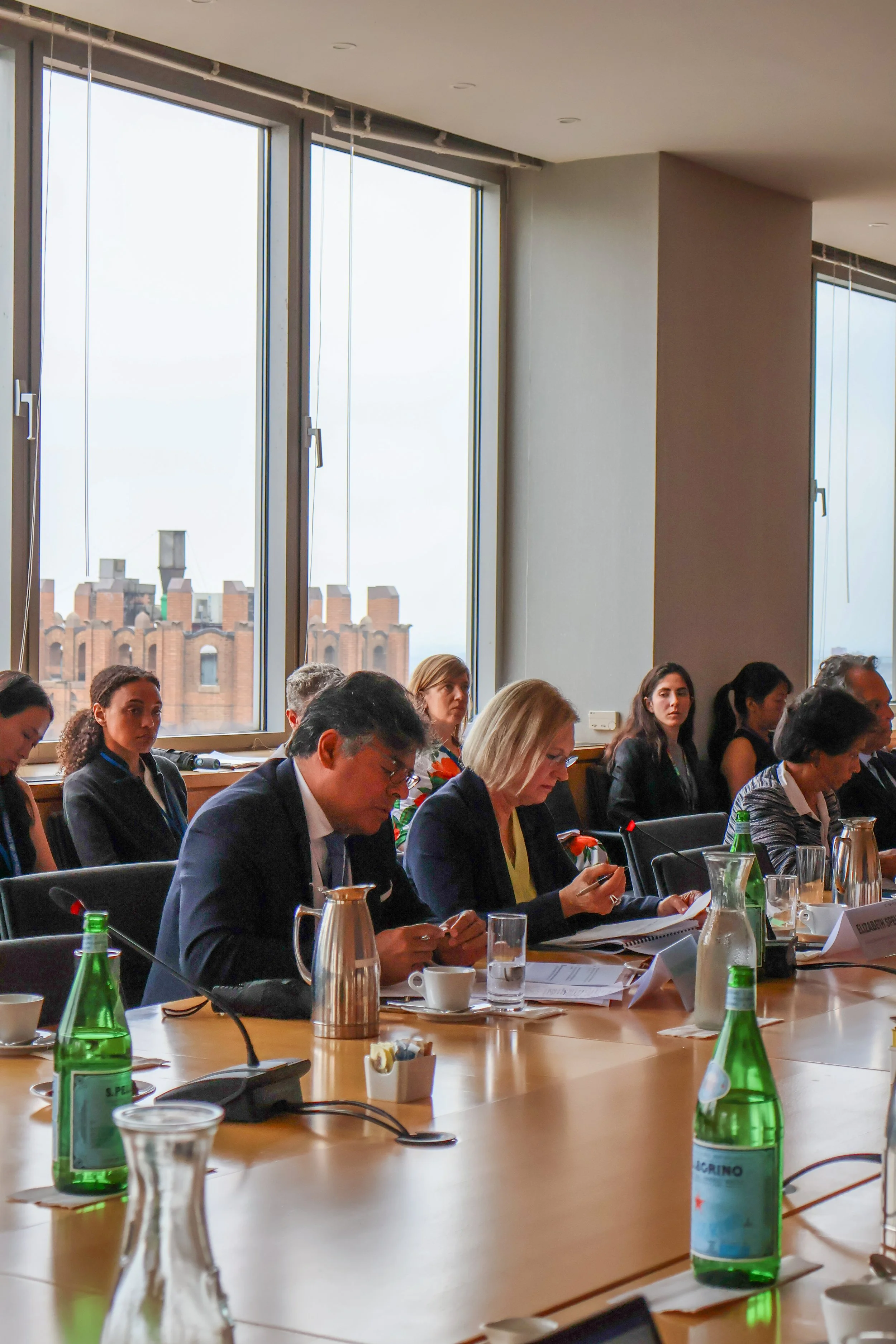 People sitting at a conference table in a room with large windows, engaged with documents and devices, with water bottles and cups on the table.