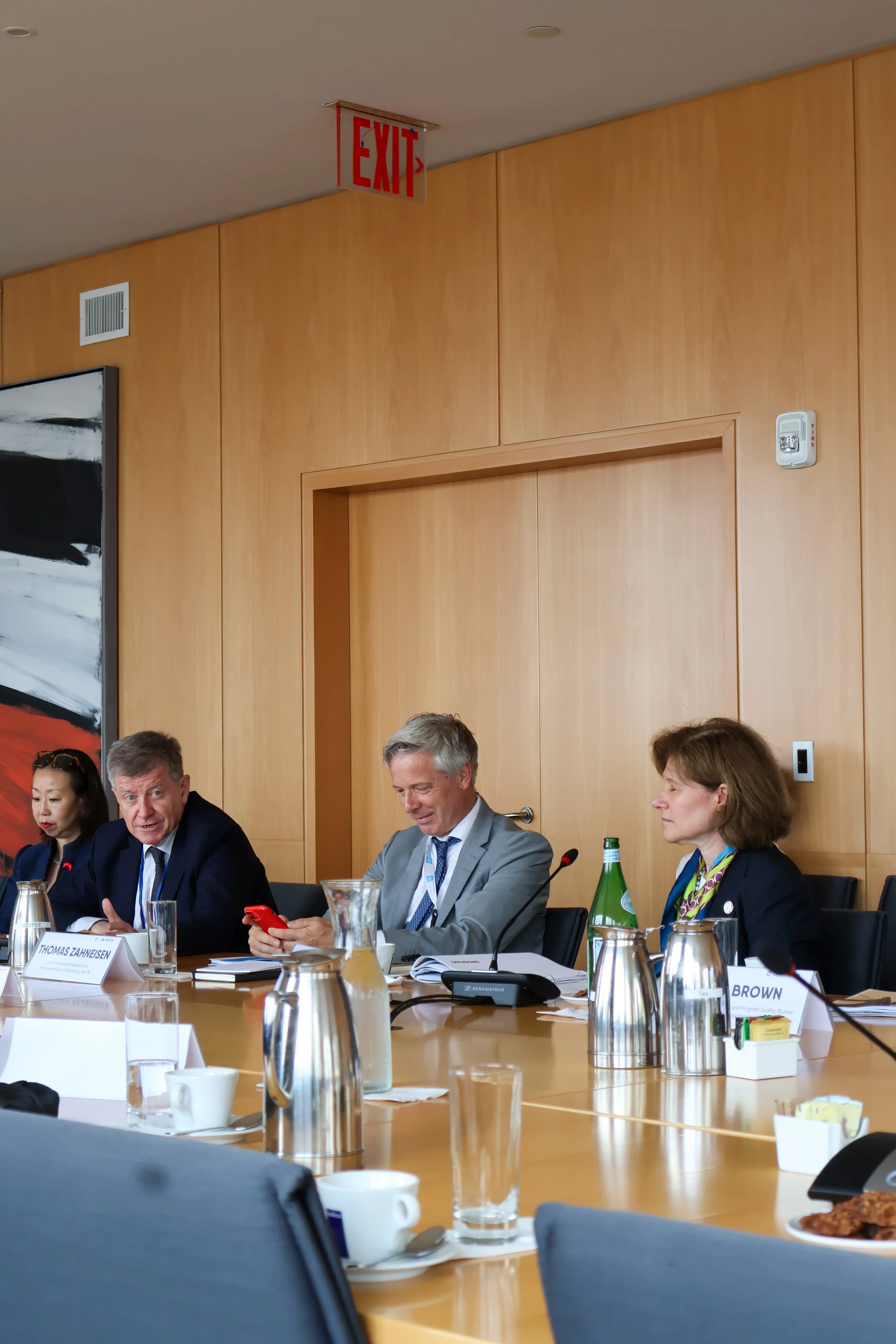 Four people are seated at a conference table in a professional setting. There are cups, glasses, water bottles, and papers on the table. The background features wood-paneled walls and a red EXIT sign hanging from the ceiling.
