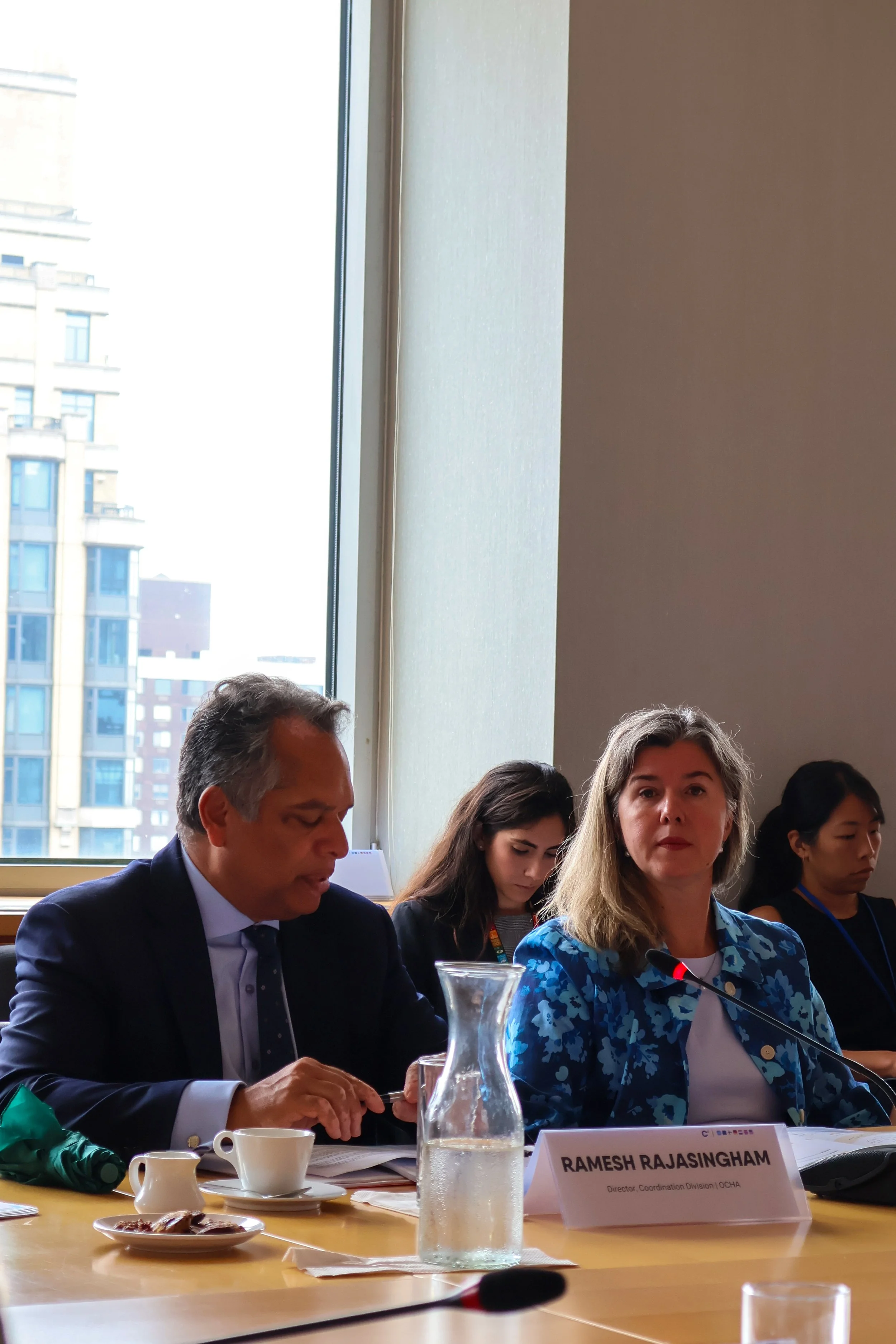 A group of people sitting at a conference table with a large window behind them, showing tall buildings outside. One woman, identified as Ramesh Rajasingham, is speaking into a microphone. There are cups, a water pitcher, and documents on the table.