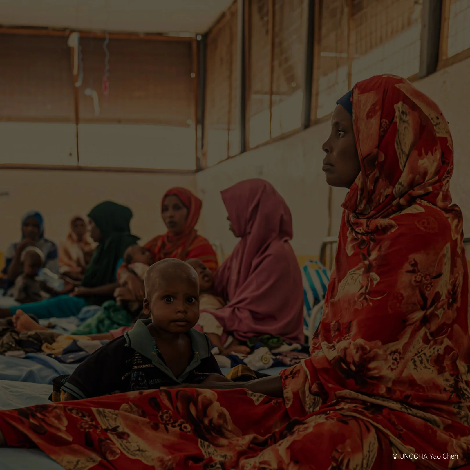 Group of women and children sitting on a hospital bed in a makeshift medical facility with wooden walls.