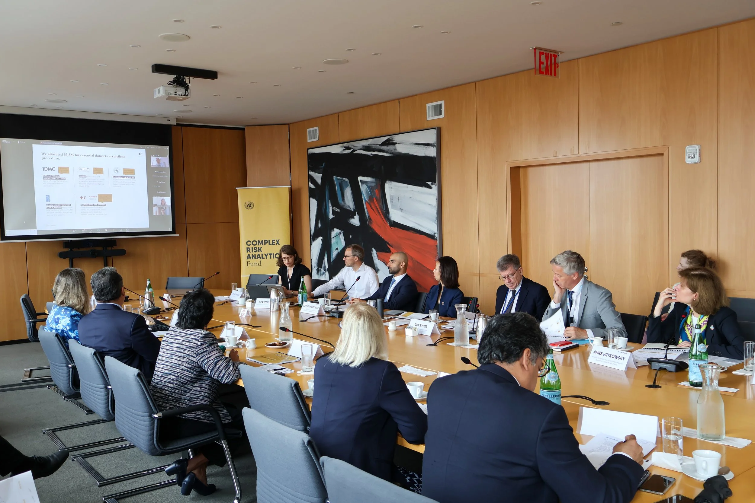 A conference room with people seated around a long table, watching a presentation on a large screen.
