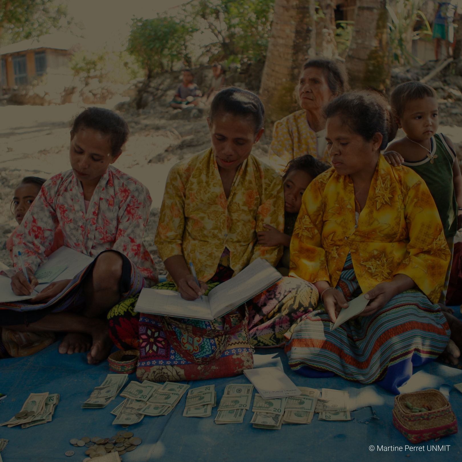 Group of women and children sitting on a blue tarp outdoors, counting and organizing US dollar bills and coins.