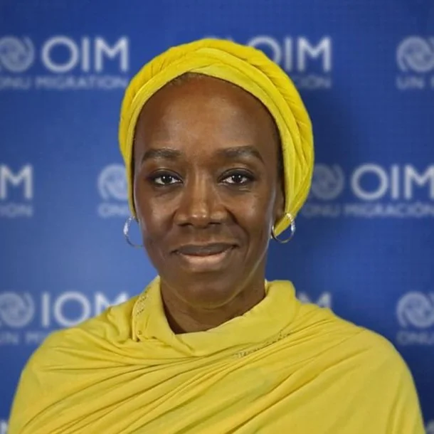 A woman wearing a yellow headwrap and matching yellow shawl standing in front of a blue background with the UN and OIM logos.