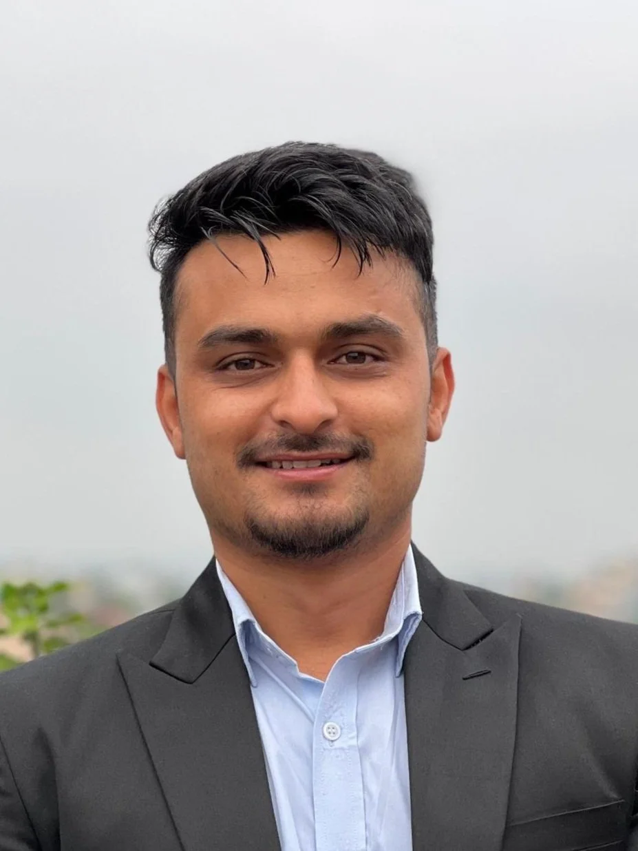 A man with short dark hair and a goatee, wearing a black blazer and light blue collared shirt, smiling outdoors on a cloudy day.
