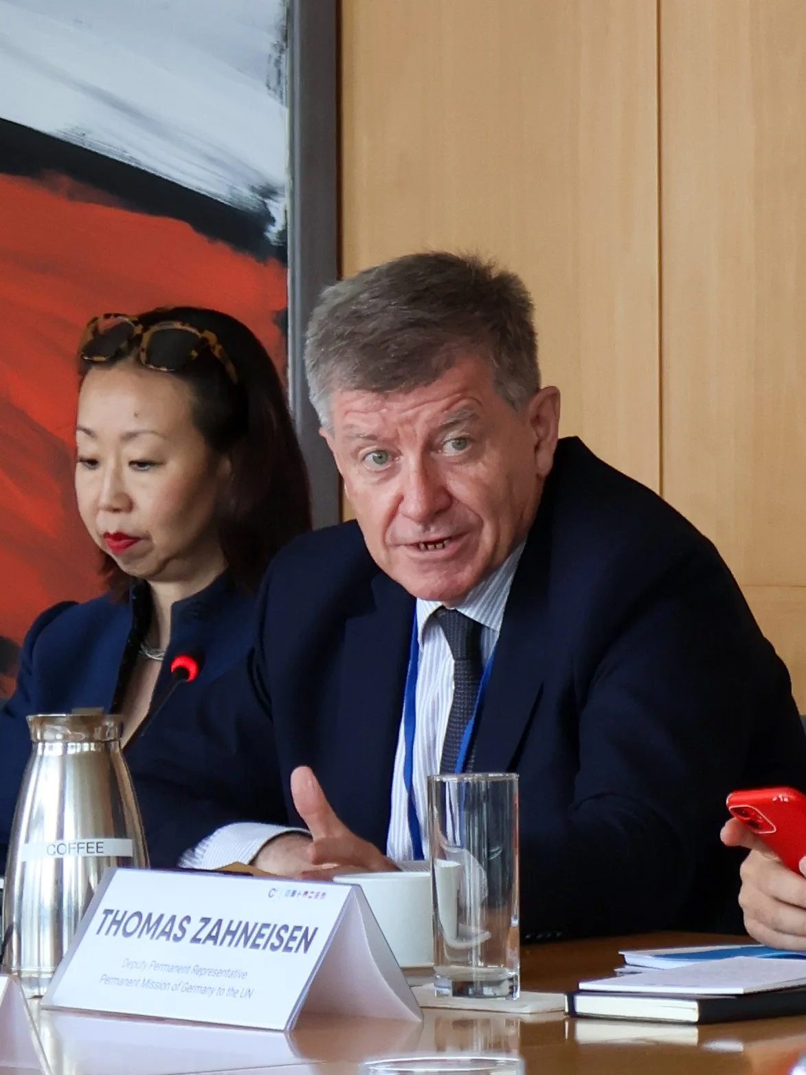 Man with gray hair in a suit speaking at a conference table, with nameplate reading 'Thomas Zahneisen', next to a woman with sunglasses on her head, and various documents and glassware on the table.