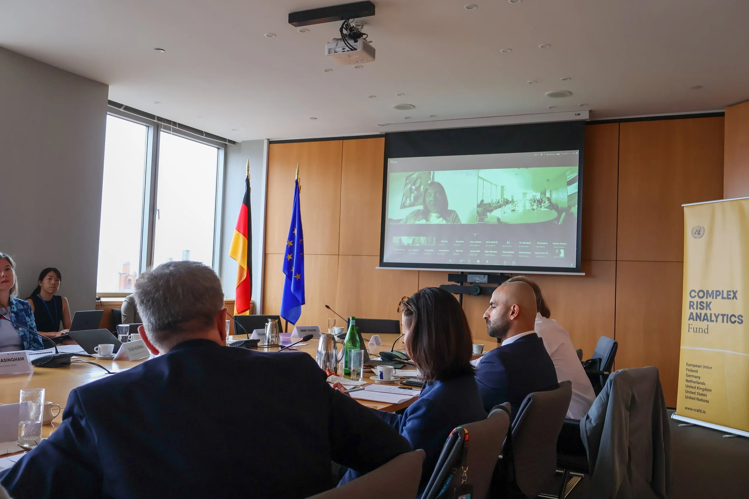 People attending a conference or meeting in a room with German and European Union flags. They are watching a video on a large screen, with laptops and papers on the table.
