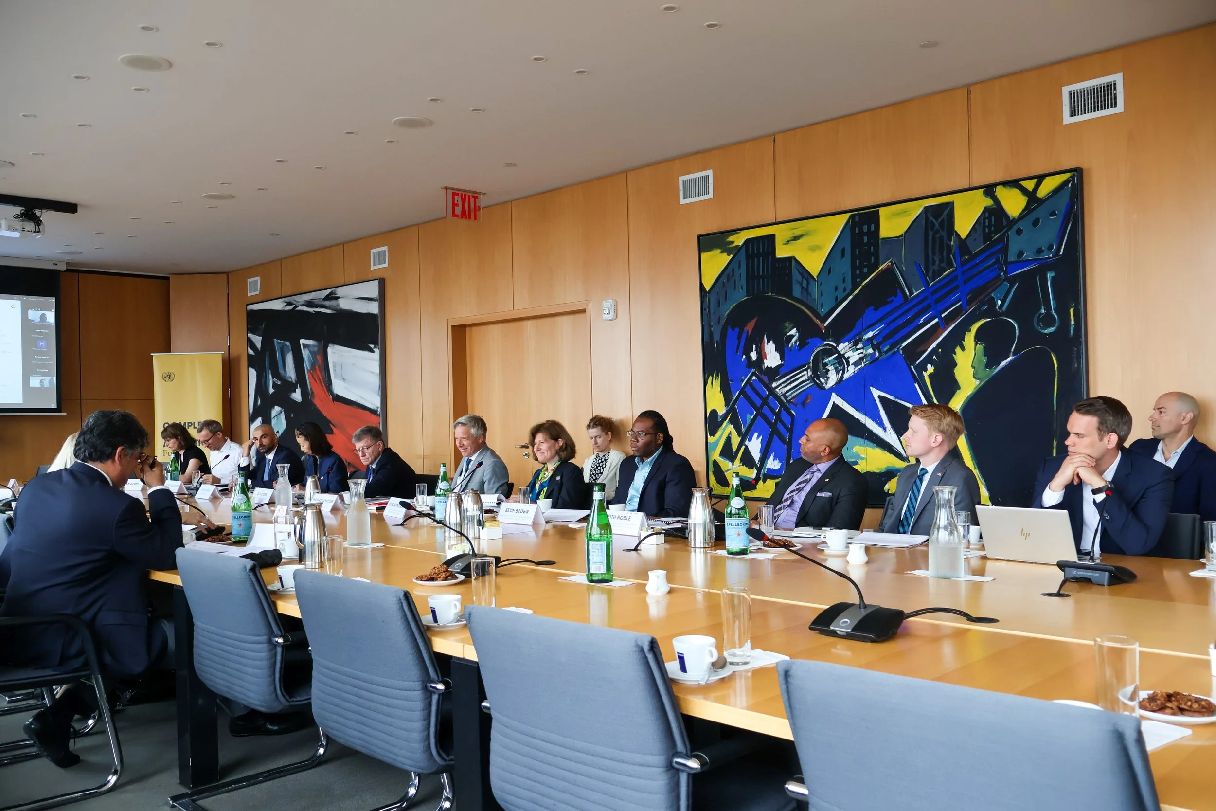 A diverse group of business professionals seated at a long conference table in a modern meeting room, with large abstract artwork on the wall behind them.