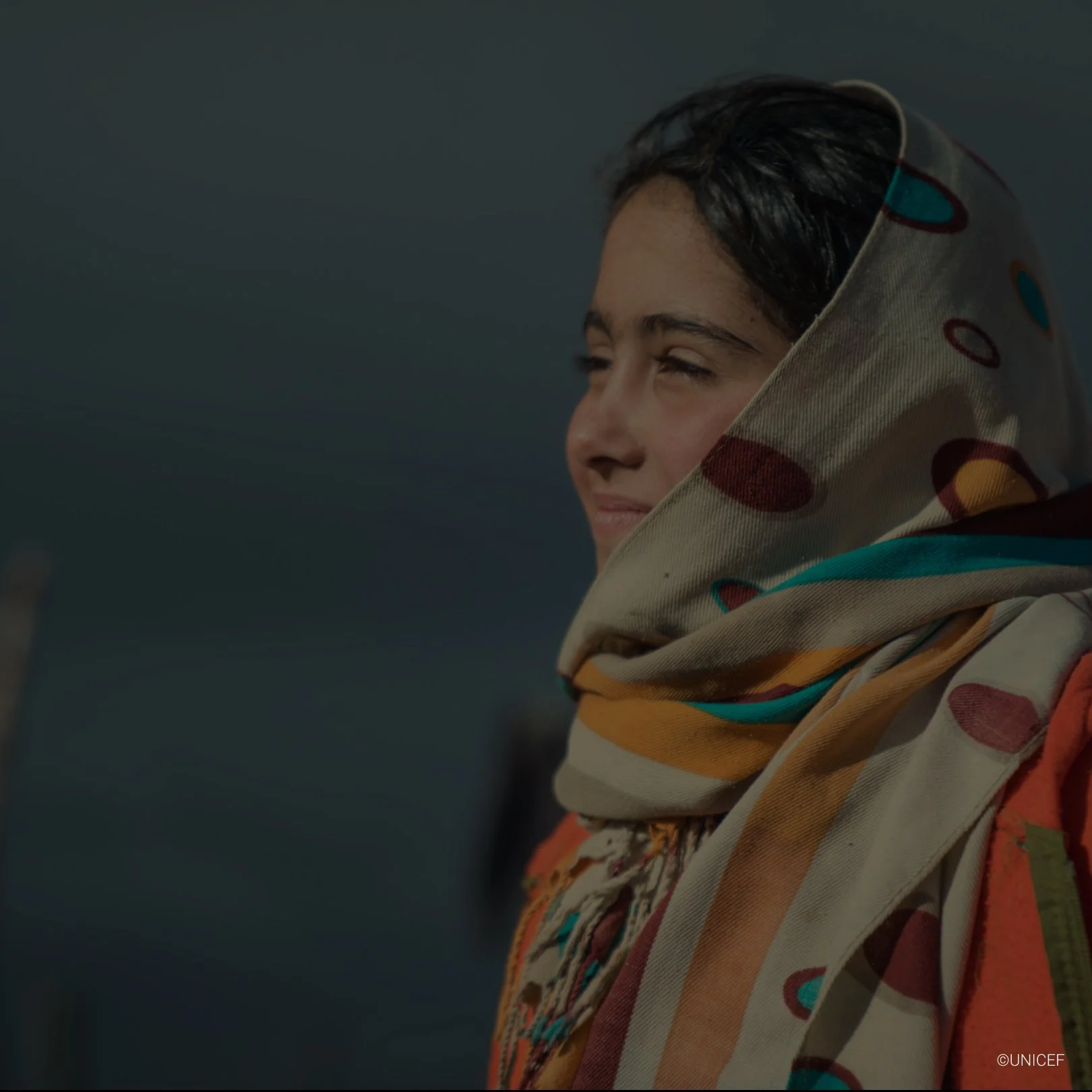 A young girl with dark hair, wrapped in a colorful patterned scarf, smiling and squinting against sunlight.