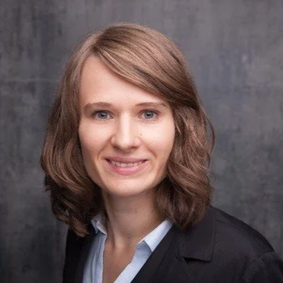Portrait of a smiling woman with shoulder-length brown hair wearing a black blazer and light blue shirt against a gray background.