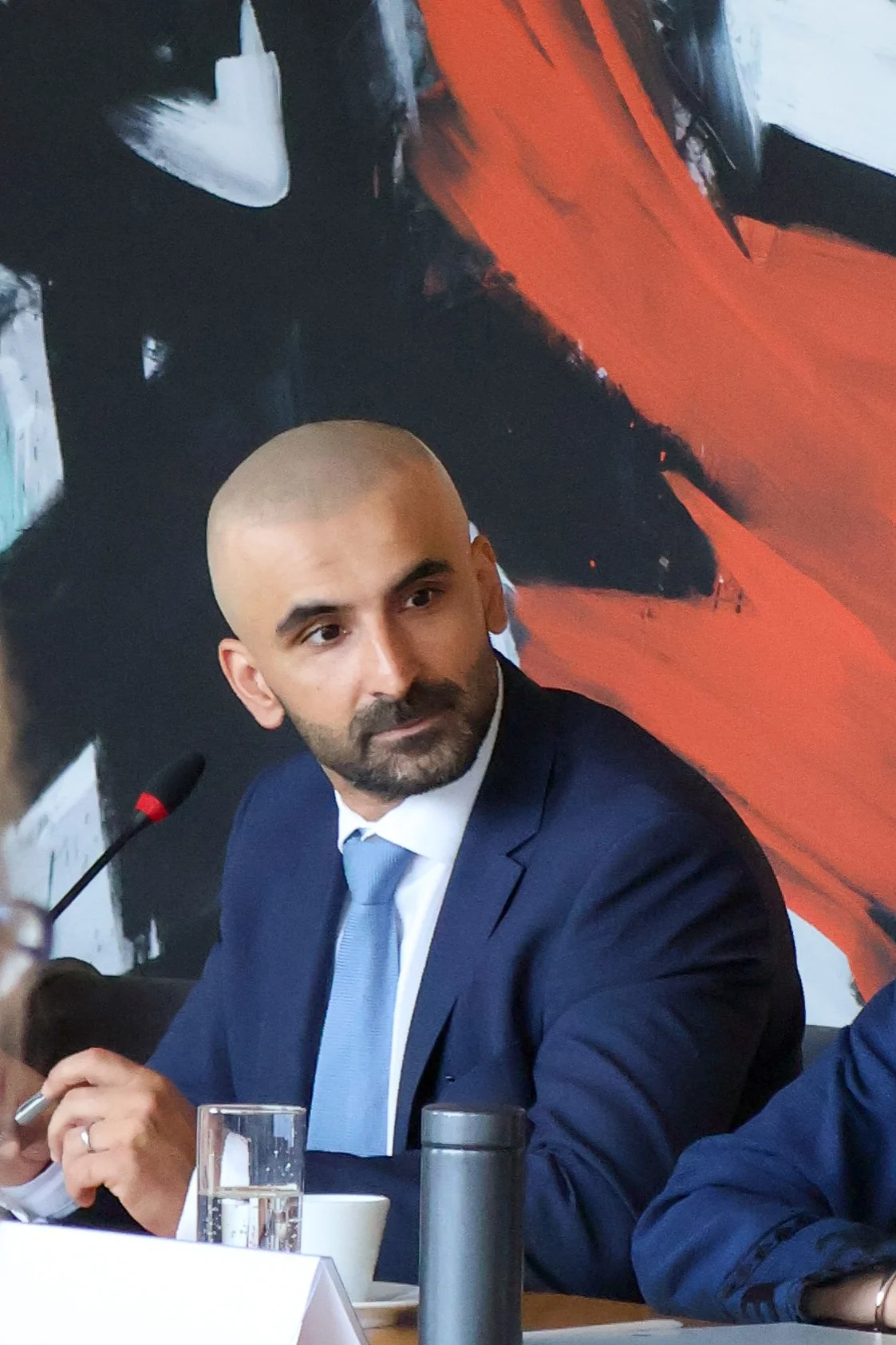 A man with a shaved head and beard wearing a navy suit, white shirt, and light blue tie, sitting at a table during a formal meeting or conference.