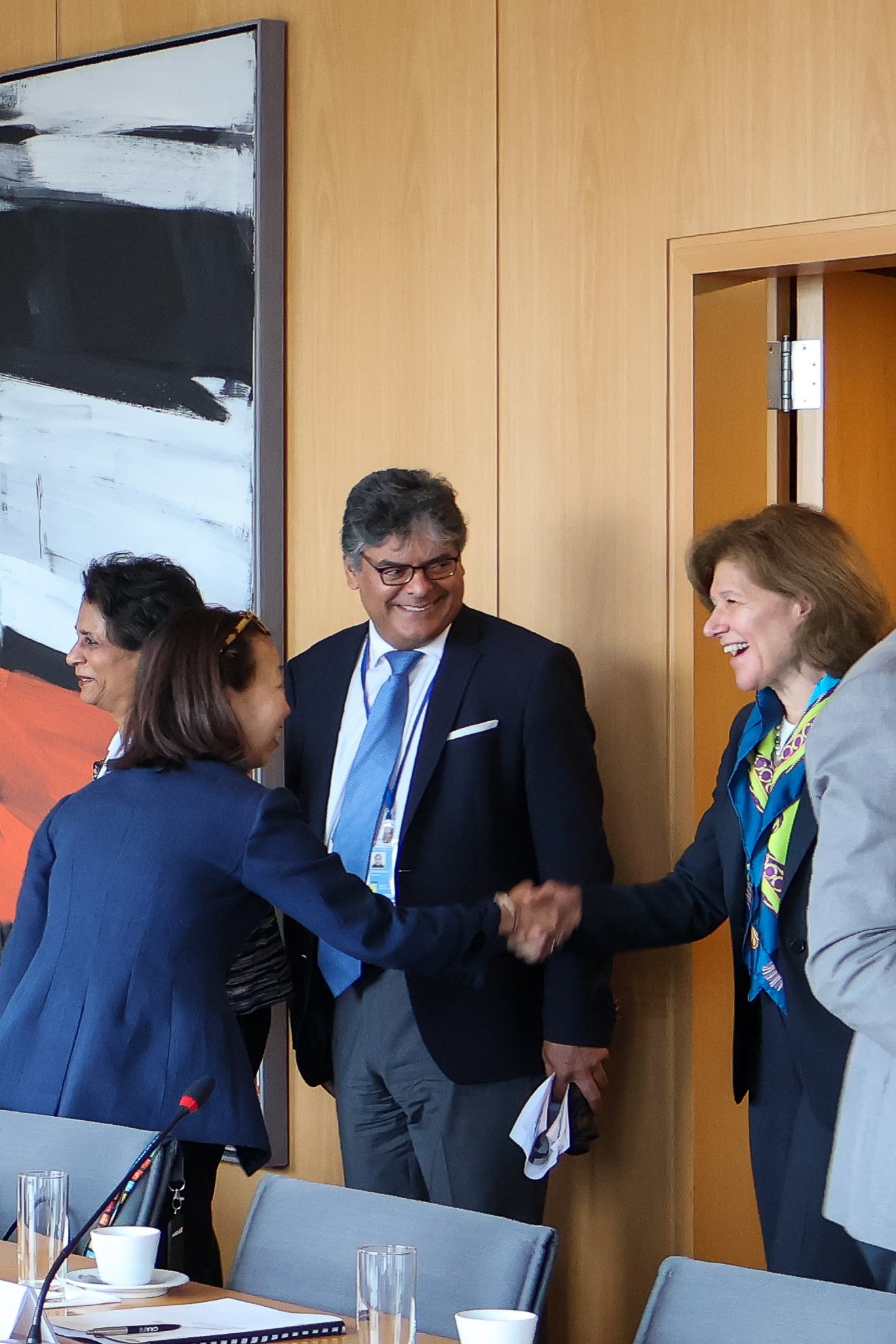 Four individuals in business attire at a meeting or conference, greeting each other with handshakes, in a room with a large abstract painting on the wall.