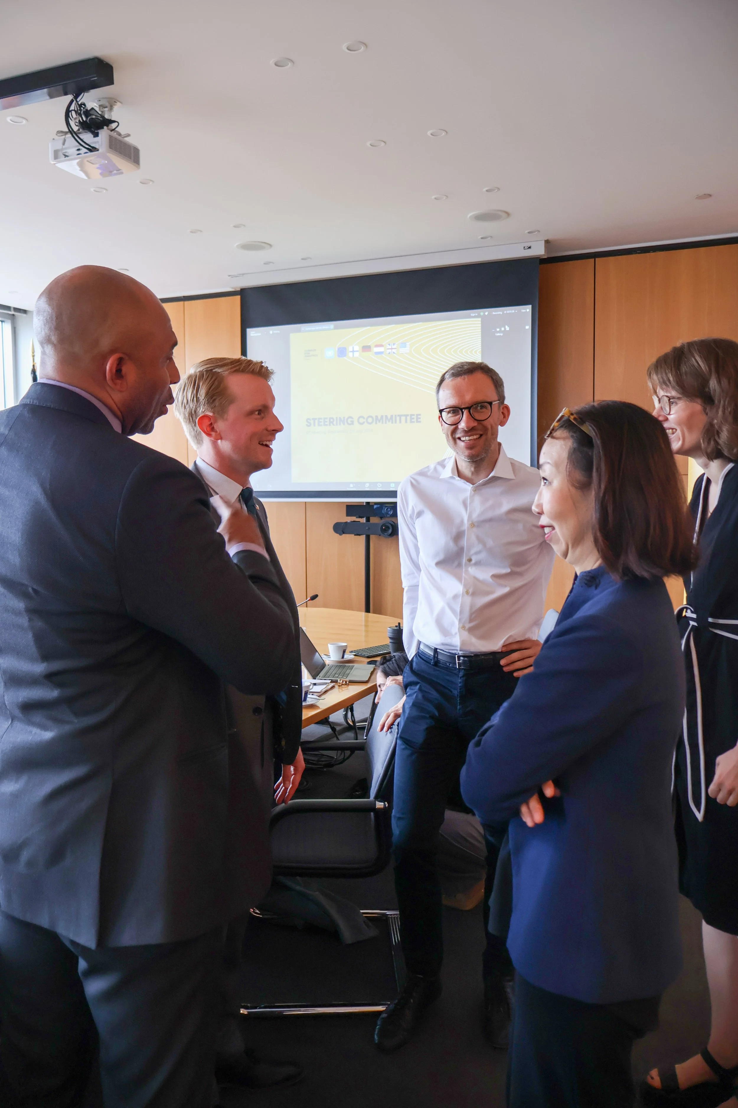 Group of six professionally dressed people standing and talking in a conference room with a presentation screen in the background.
