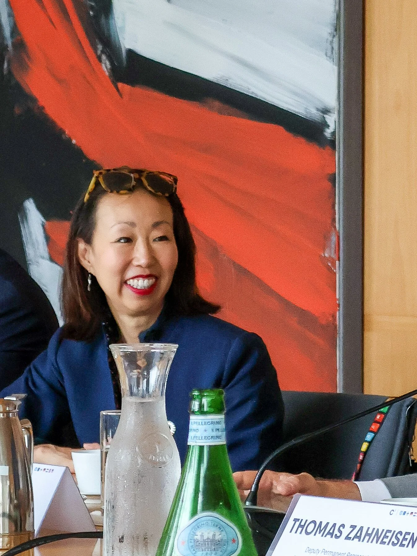 A woman with shoulder-length dark hair, wearing a blue blazer, smiling and sitting at a table with a glass pitcher and a green glass bottle, in front of a large abstract painting with red, black, white, and gray colors.