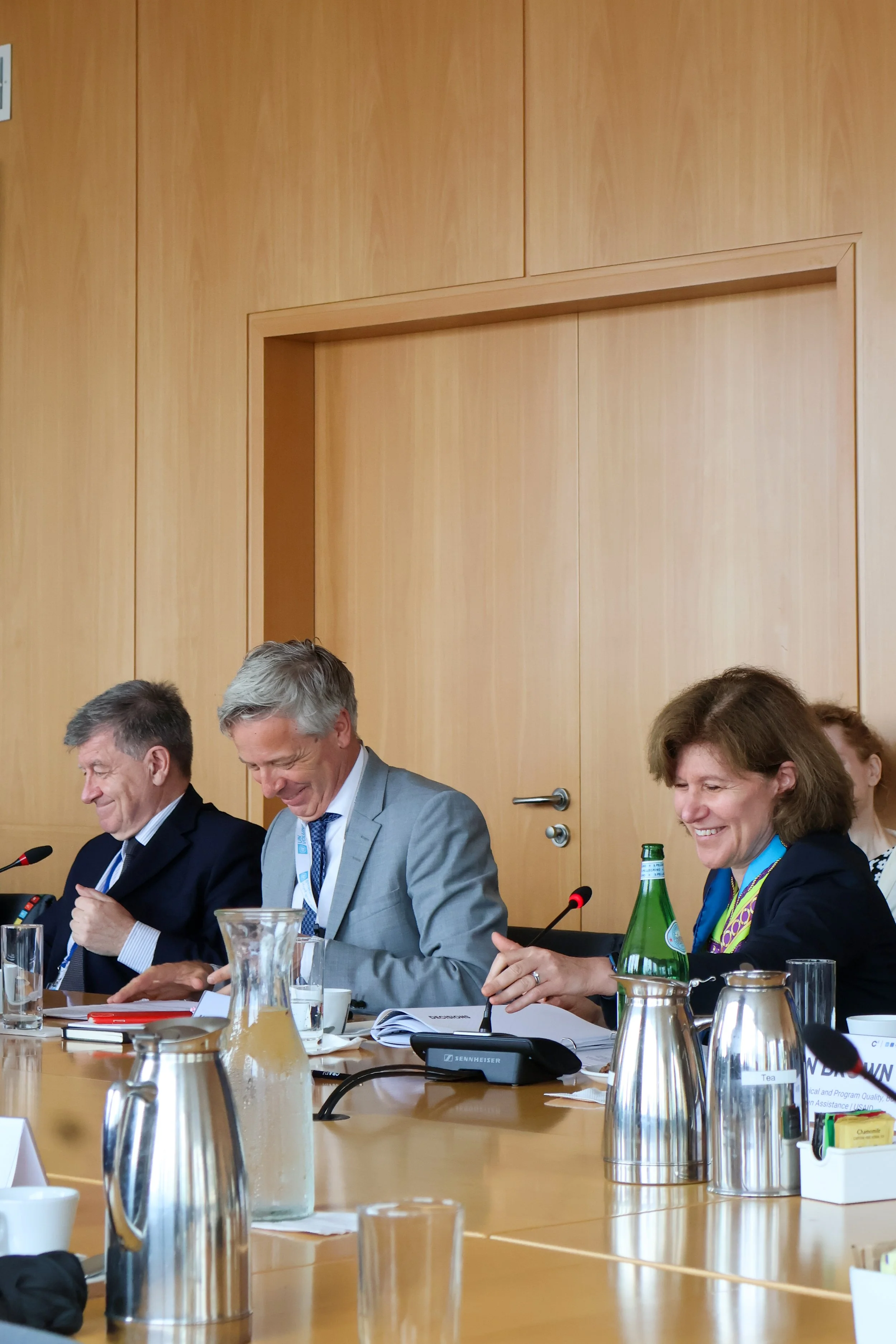 Three people sitting at a conference table, smiling and engaging during a meeting, with microphones, water bottles, and notebooks on the table.