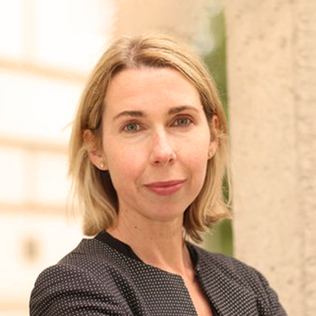 A woman with blonde hair and blue eyes, wearing a black polka dot blazer, standing outdoors near a stone column.