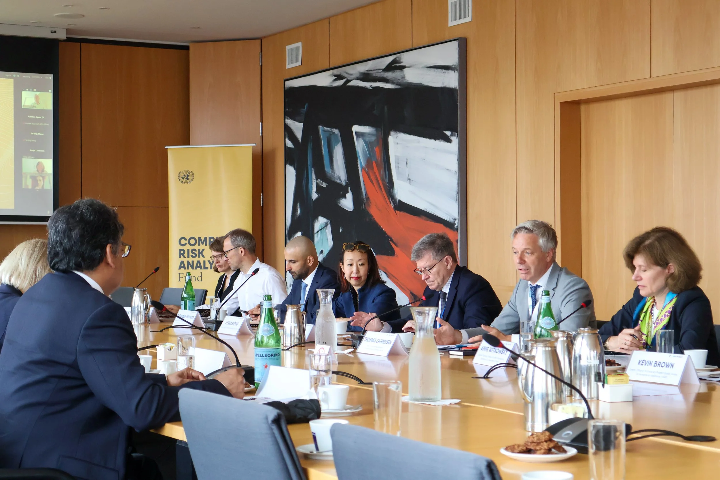 A group of business professionals sitting at a conference table during a meeting or discussion, with bottles of water, glasses, and papers on the table, in a wood-paneled room with an abstract painting on the wall.