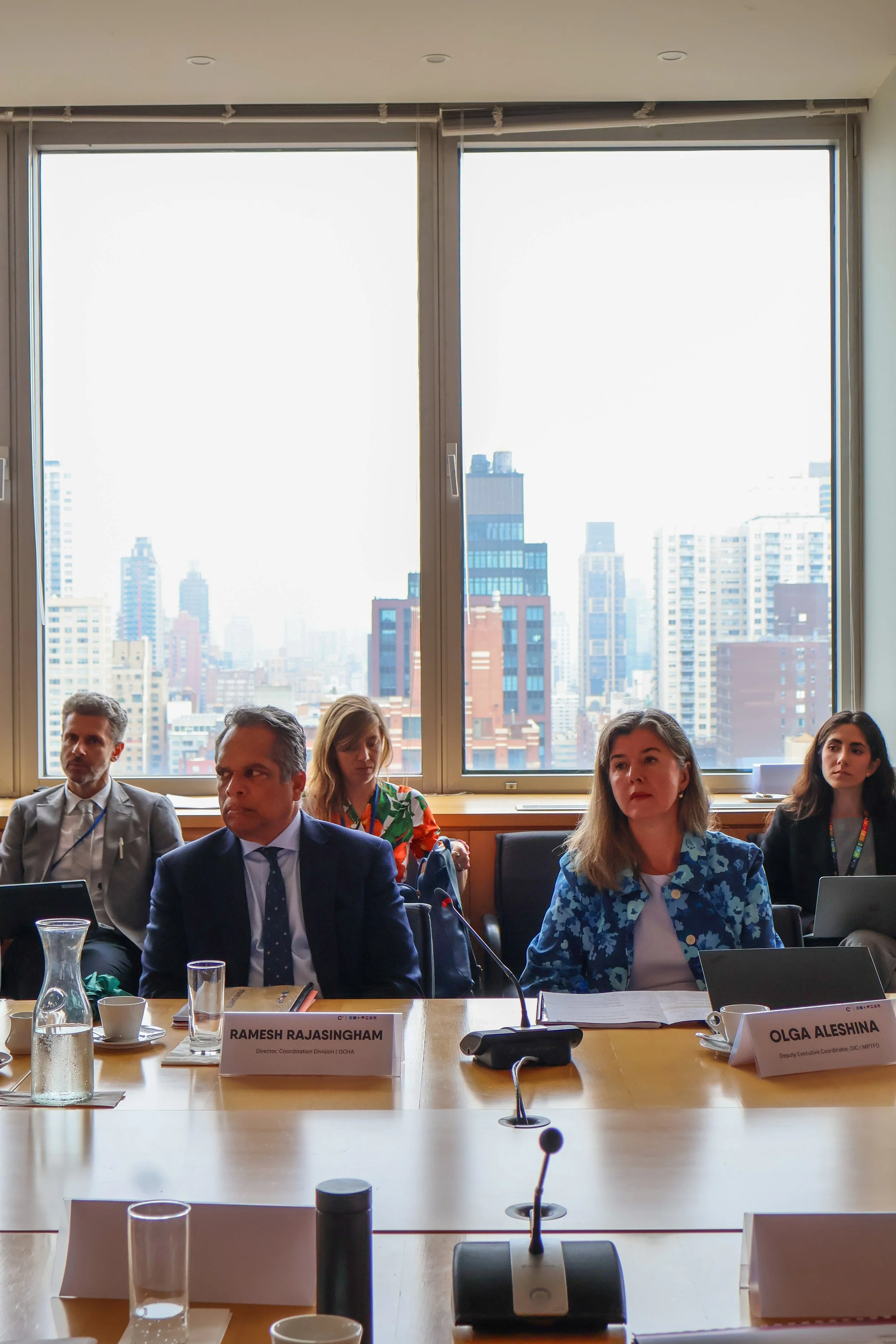 A group of five people sitting at a conference table in a high-rise office with large windows showing city skyscrapers. The two women and three men are dressed professionally, with notebooks, microphones, and nameplates in front of them.
