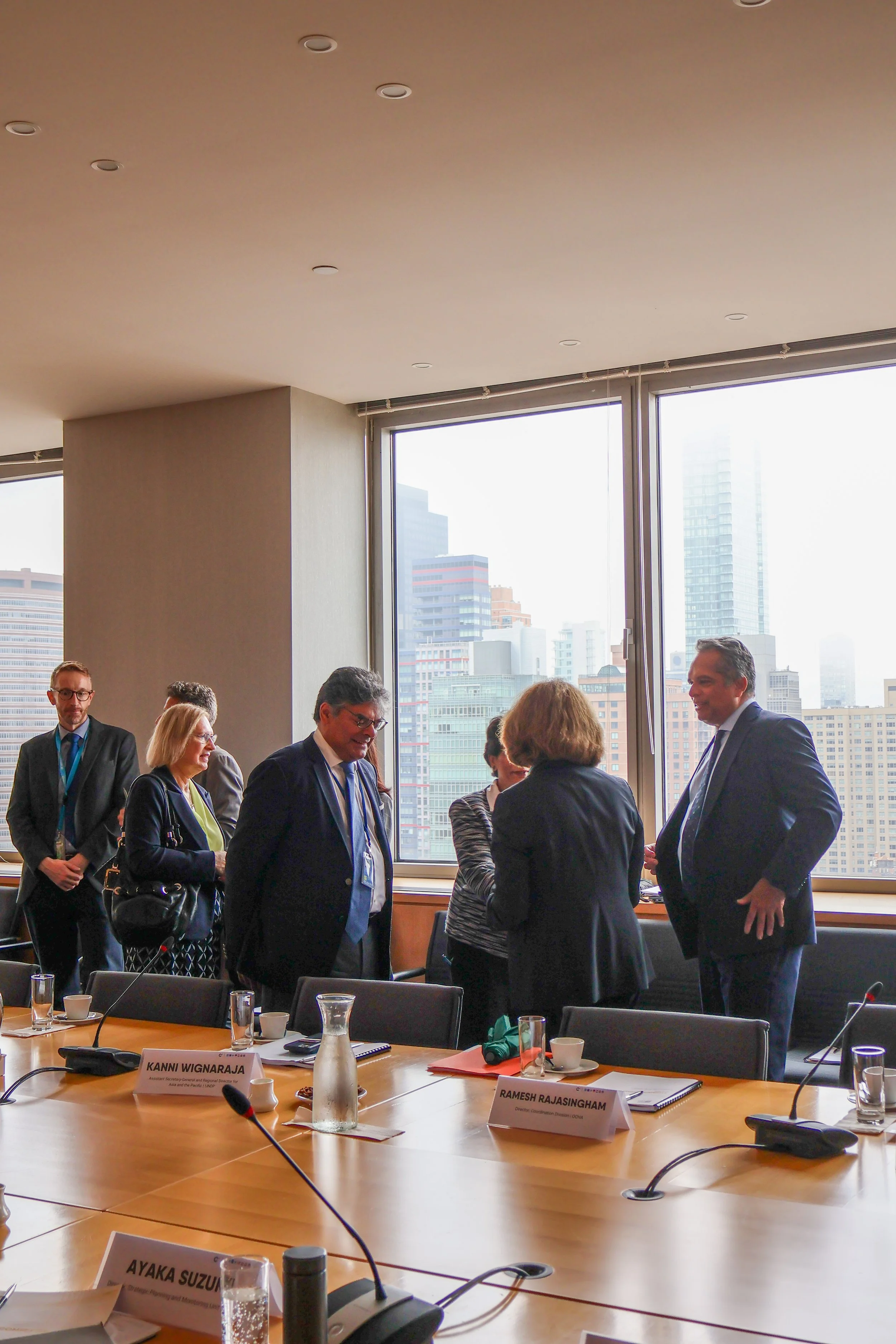 Business people engaging in conversation around a conference table in a high-rise office with city skyline view.