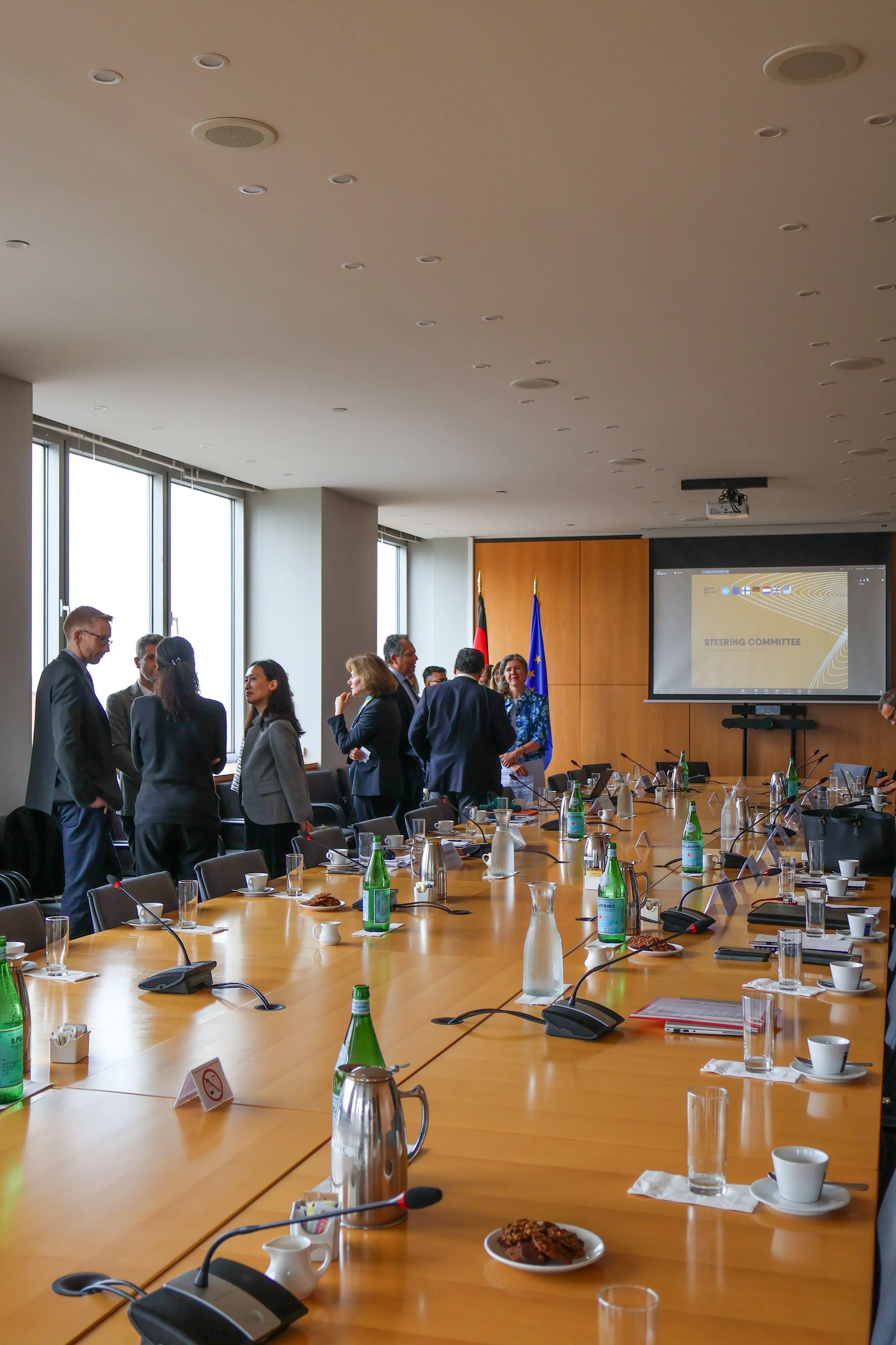 Conference room with a long table, water bottles, glasses, cups, and microphones, with people standing and talking near the windows and a presentation screen displaying 'Steering Committee.'