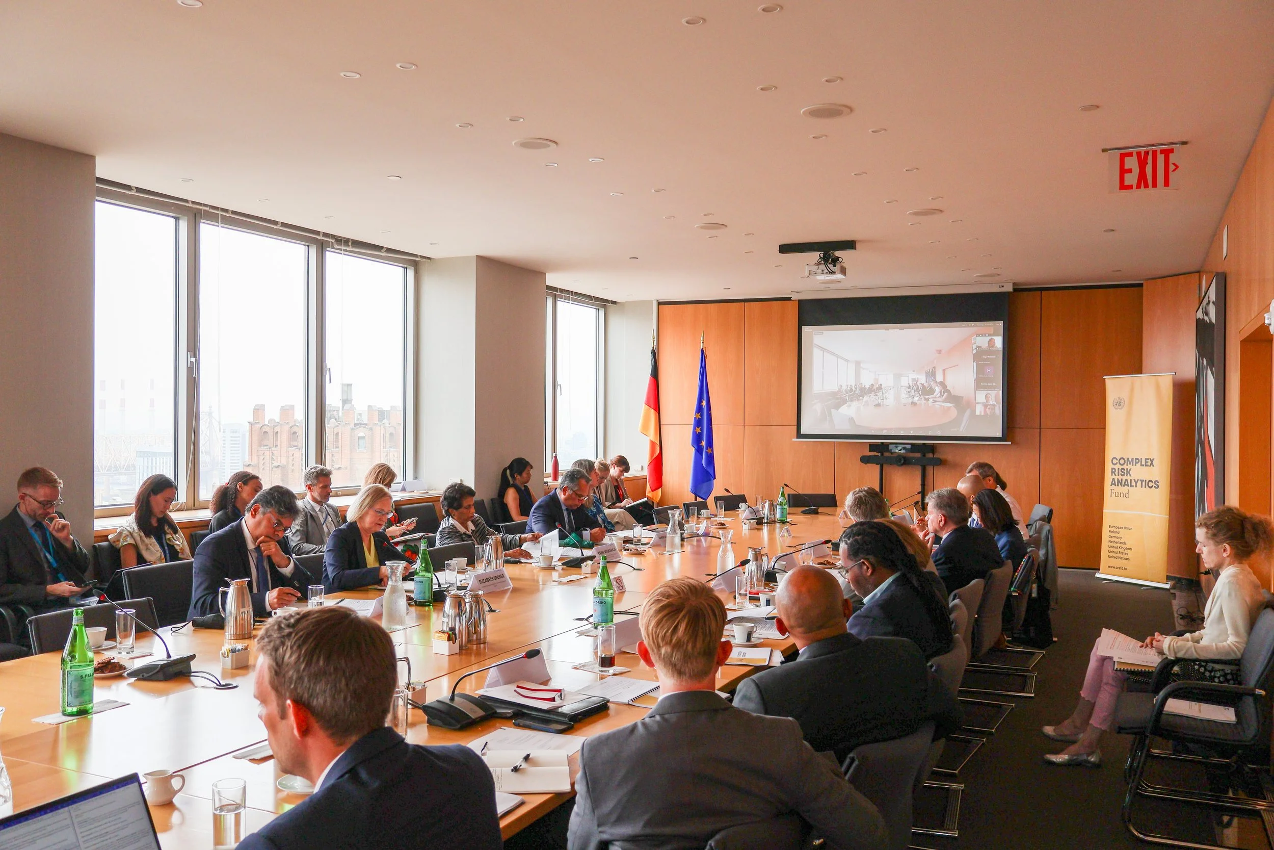 A large conference room filled with people seated around a long table, participating in a meeting or presentation. There are two flags, the German flag and the European Union flag, positioned at the front of the room. A large screen displays a video 