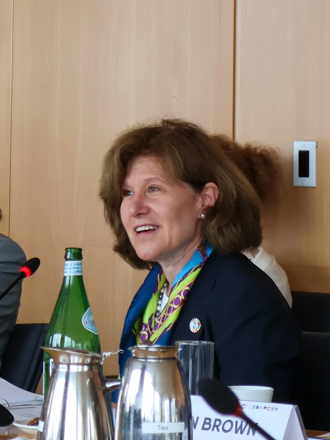 A woman with brown hair smiling, sitting at a conference table with a green glass water bottle and metal pitchers in front of her, wearing a dark blazer and colorful scarf.