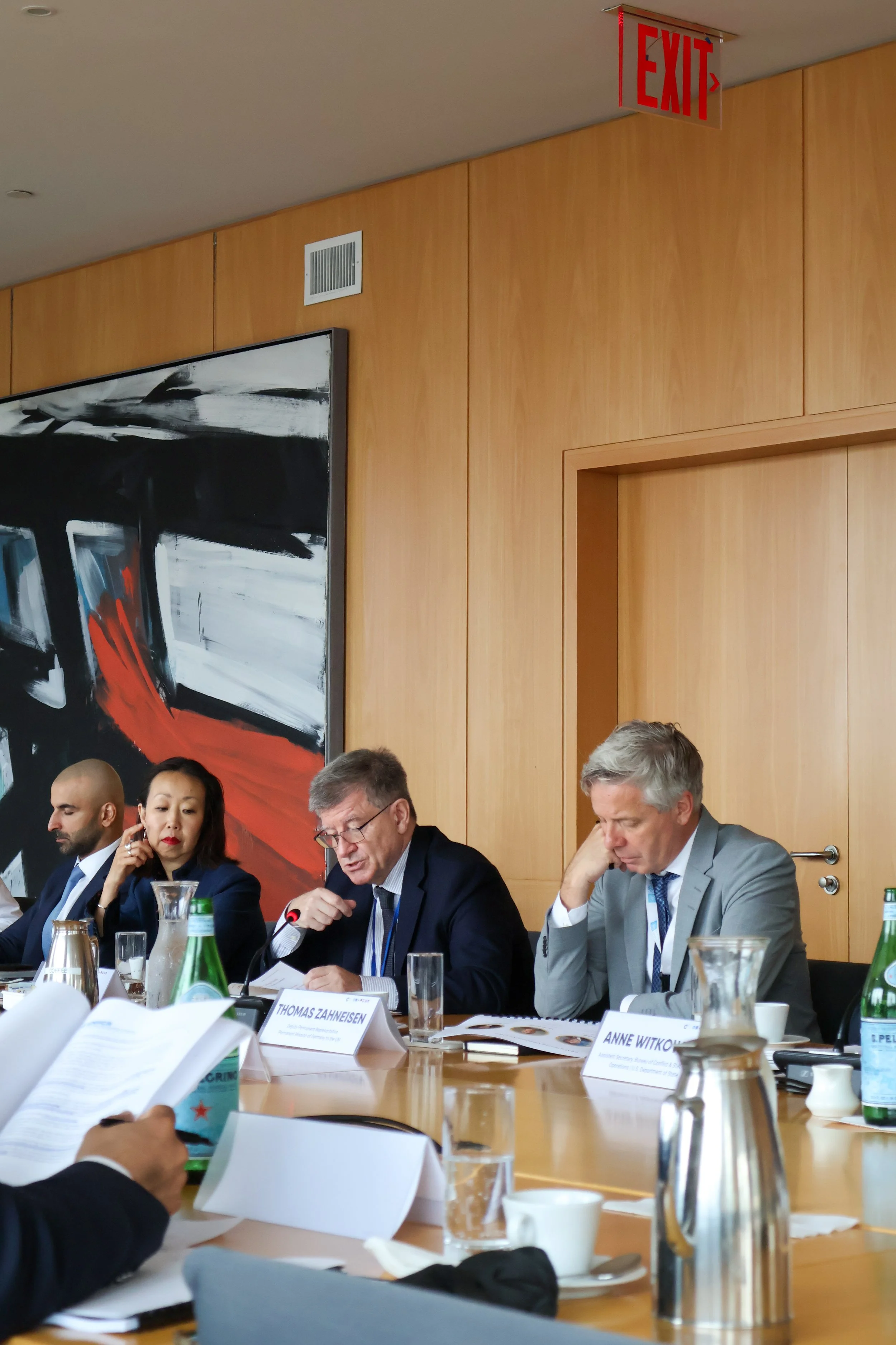 A group of professionals, including men and women, sitting at a conference table during a meeting, with documents, water bottles, and glasses on the table, in a room with a wooden wall and large abstract painting.