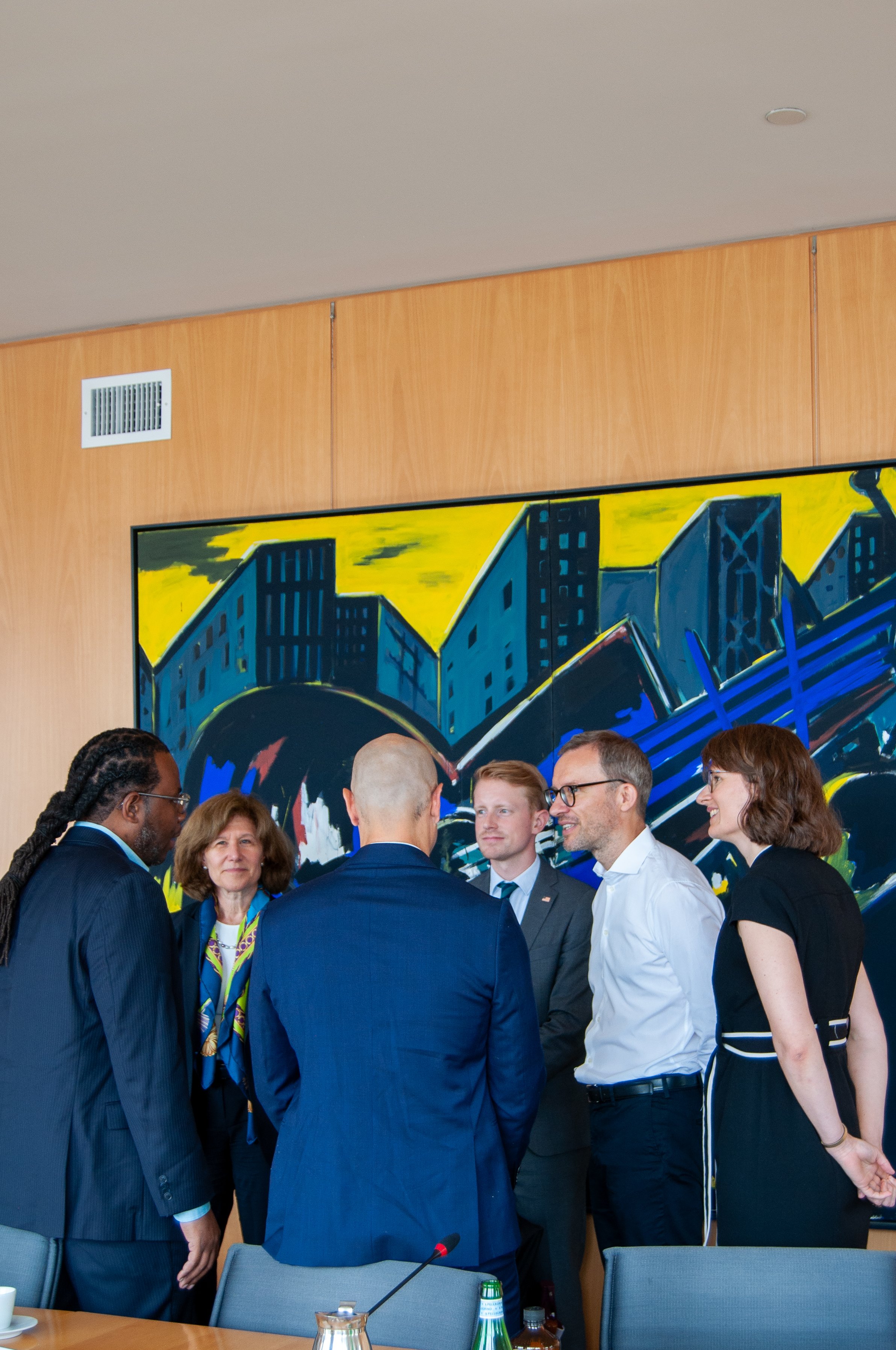 Group of six professionals having a conversation in a conference room with a colorful abstract cityscape painting on the wall behind them.