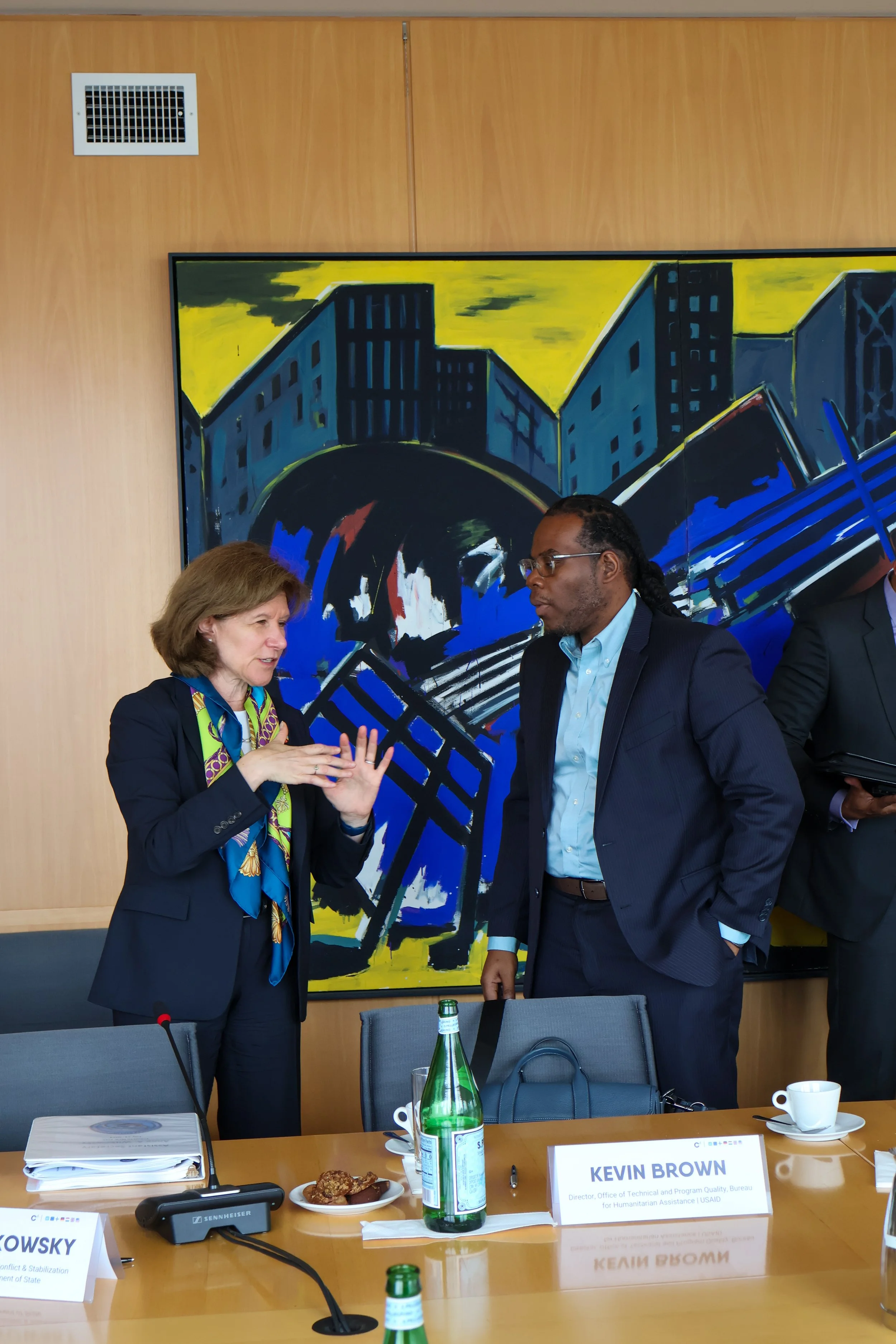 Two professionals, a woman and a man, engaged in conversation at a conference table. The woman is using hand gestures, wearing a dark blazer and colorful scarf. The man is dressed in a dark suit and light blue shirt. Behind them is a large, abstract 