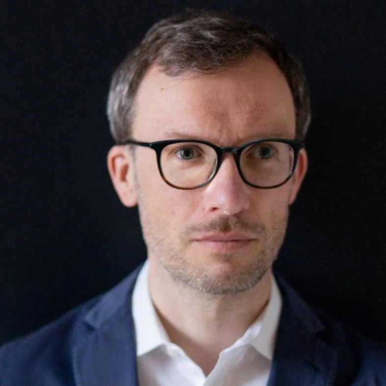A man with glasses and brown hair wearing a suit, posing against a black background.