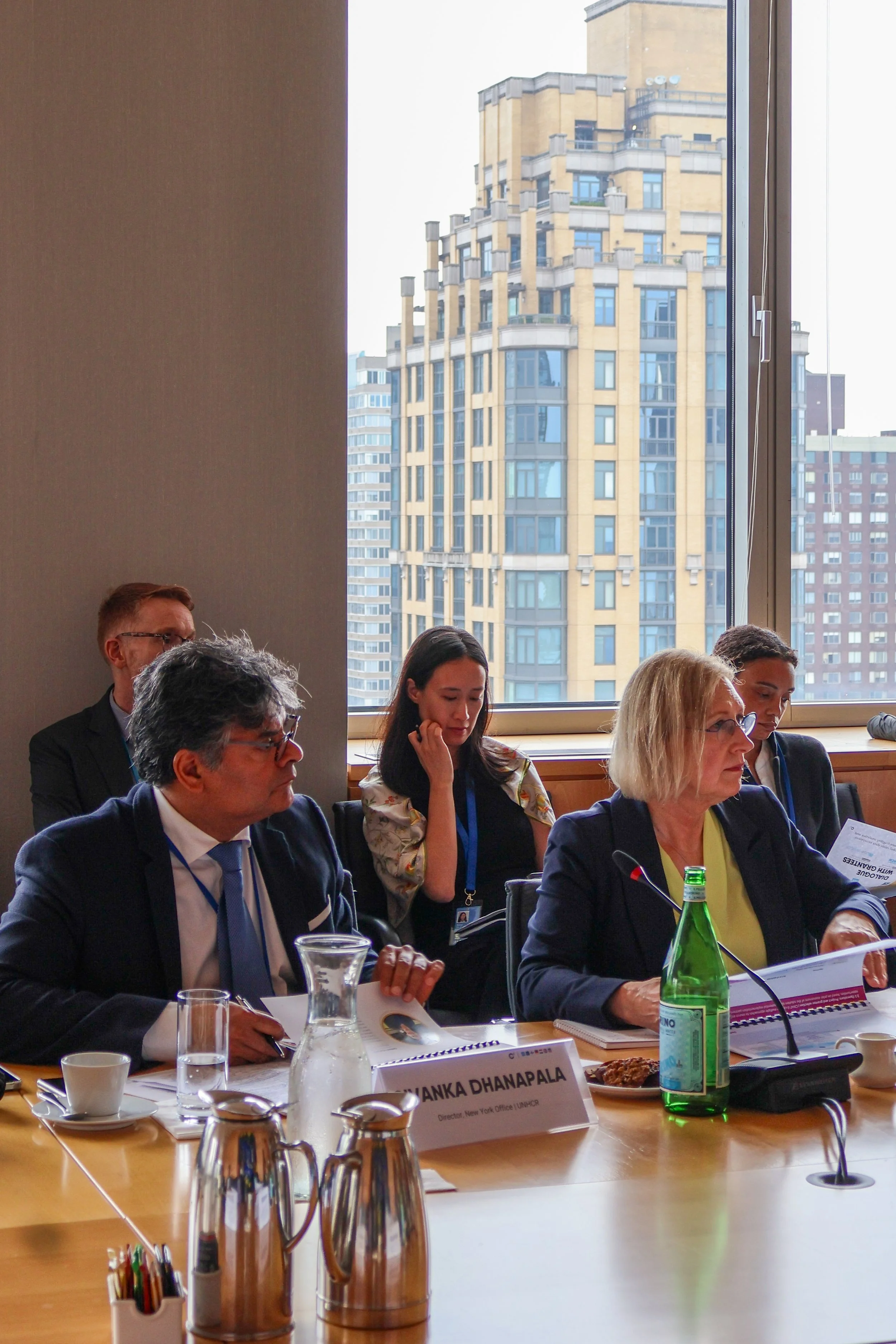 A group of diverse professionals sitting at a conference table during a meeting, with a cityscape of tall buildings visible through a large window behind them.