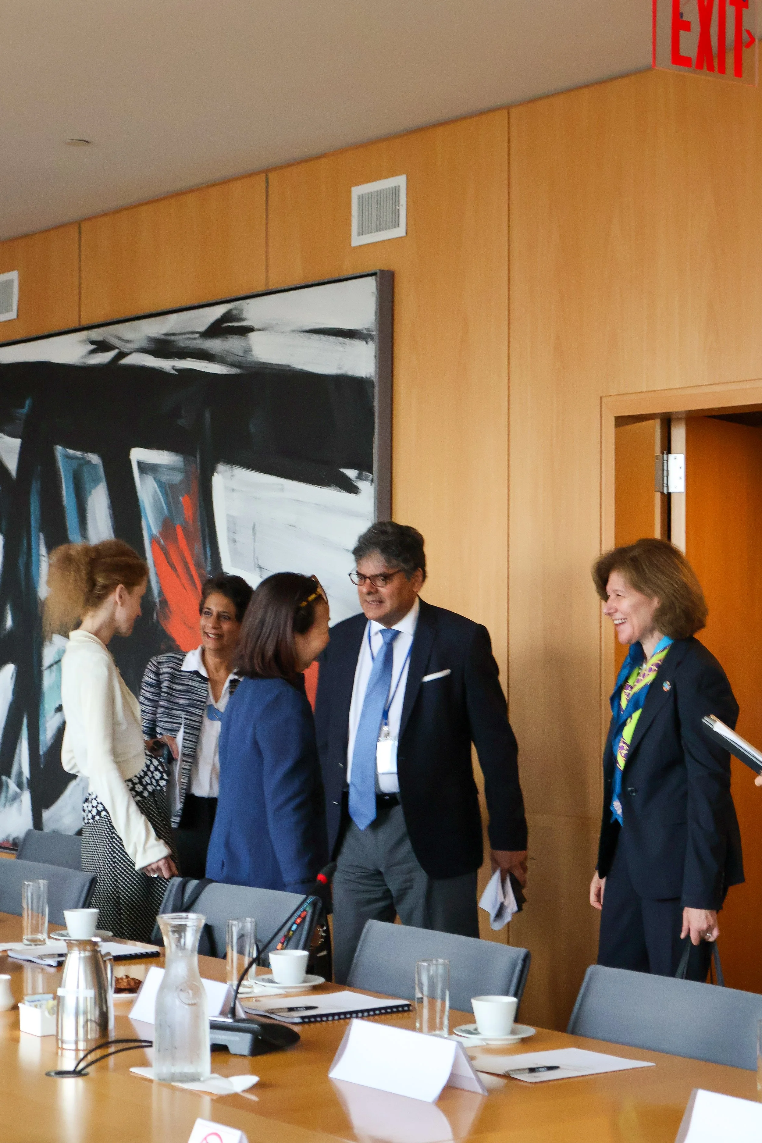 Group of five people, three women and two men, engaged in conversation in a conference room with a wood-paneled wall and abstract painting, with a long table set with glasses, cups, and notebooks.