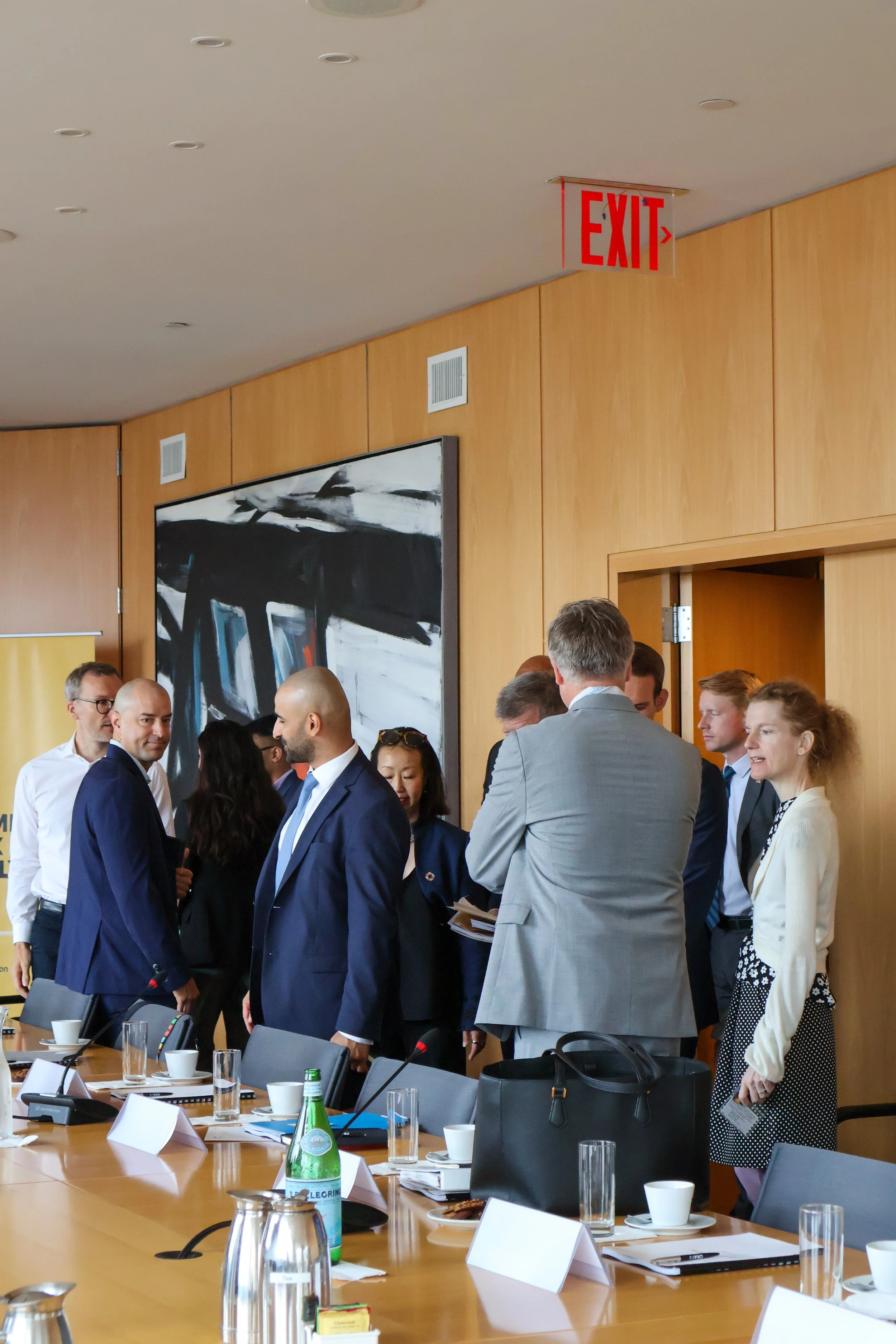 A group of business professionals in suits gathered in a meeting room, standing and talking near a conference table with documents, water bottles, and cups.