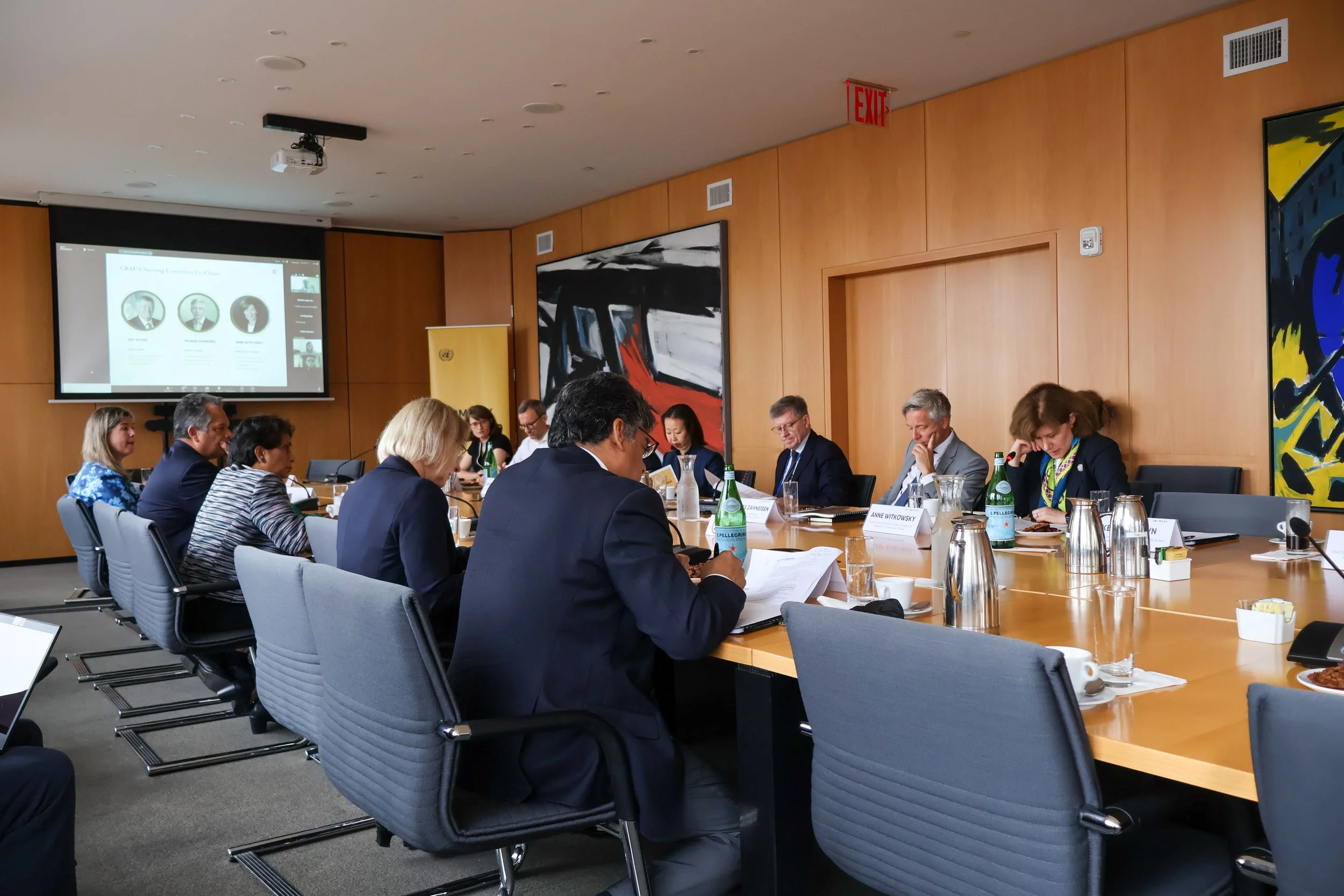 Business meeting in a conference room with attendees seated around a large table, a presentation screen on the wall, and bottled water on the table.