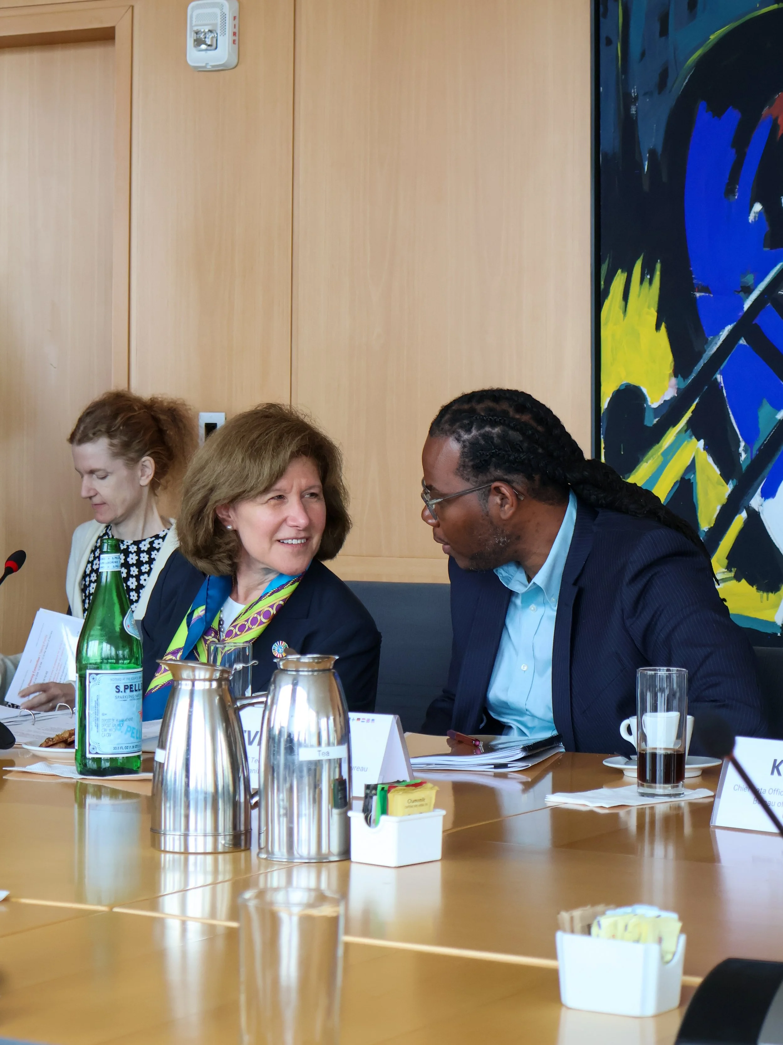 Two women and a man sitting at a conference table, engaged in conversation. The woman on the left has blonde hair and is smiling, the man on the right has dreadlocks and is wearing glasses, and the woman in the background has red hair and is reading 