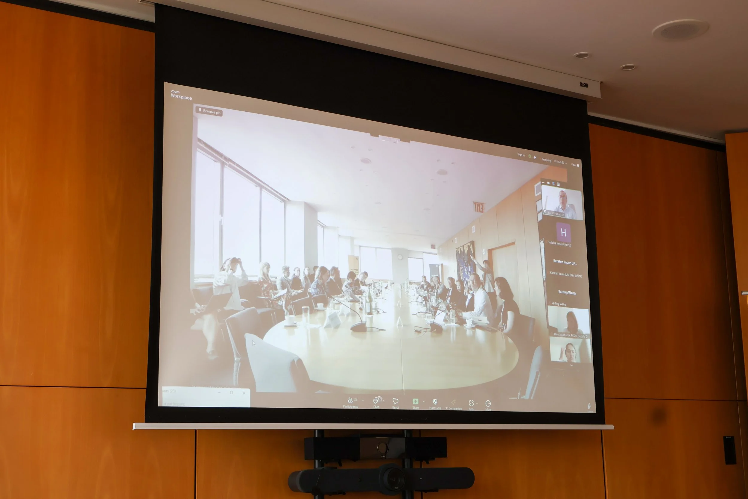 A conference room with a large round table and many people sitting around it, visible on a projection screen, during a video call.