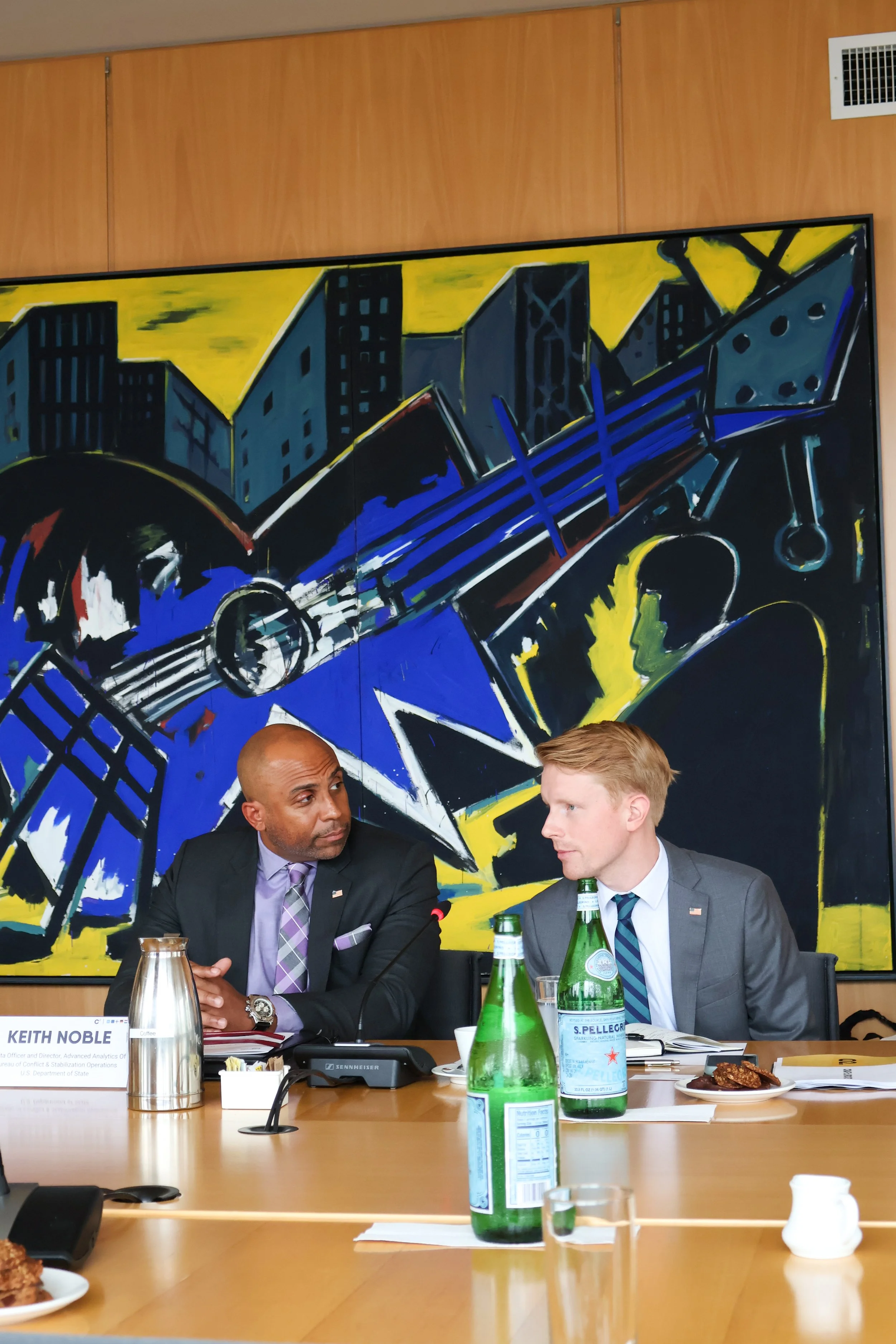 Two men in suits seated at a conference table with bottles of San Pellegrino sparkling water, a glass, and plates of food; a large colorful abstract painting of a cityscape hangs on the wall behind them.