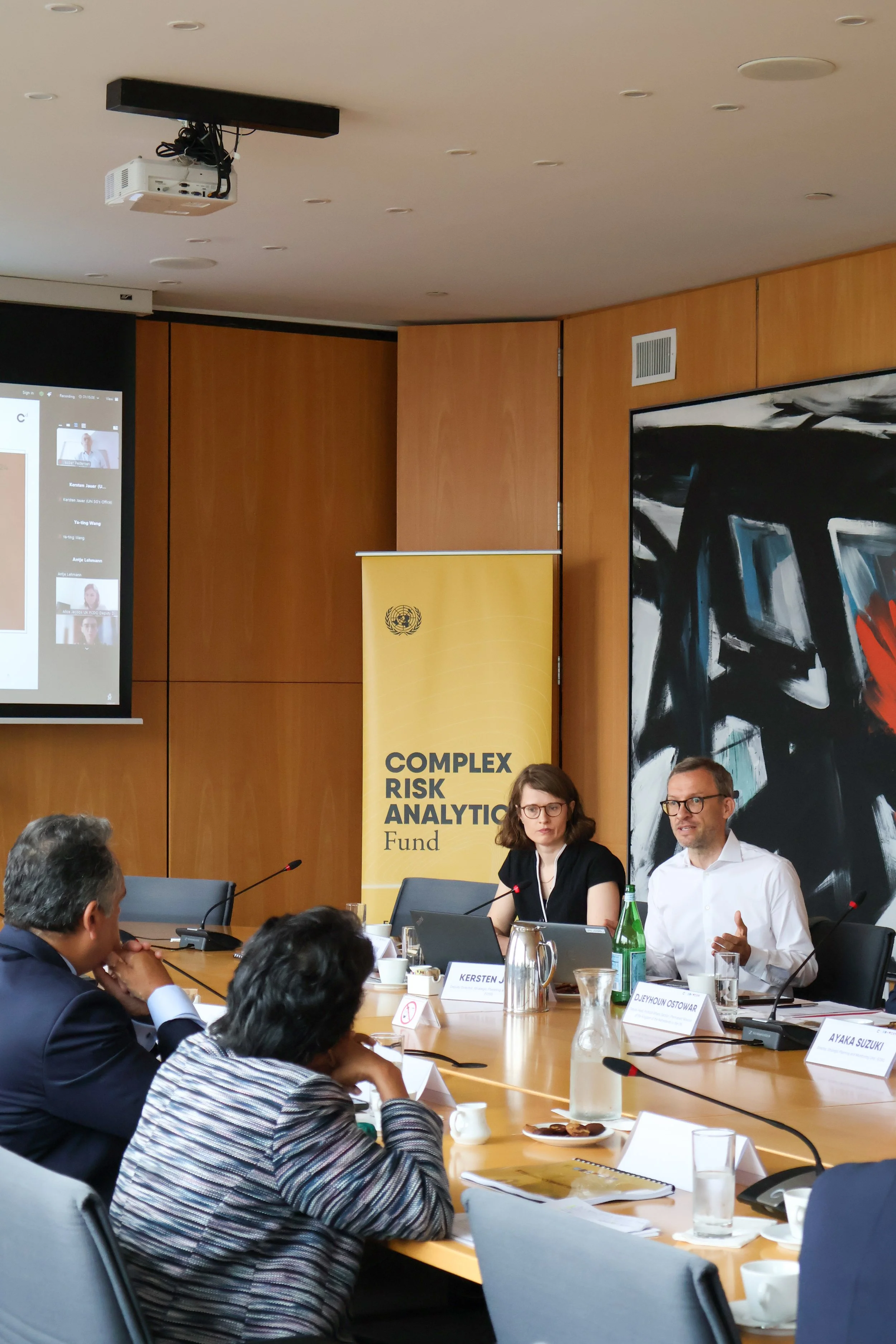 A conference room with a group of people sitting around a wooden table, some taking notes, and a woman with glasses speaking. Two people are visible in the foreground, and a yellow banner with 'COMPLEX RISK ANALYTICS Fund' stands behind the speakers.