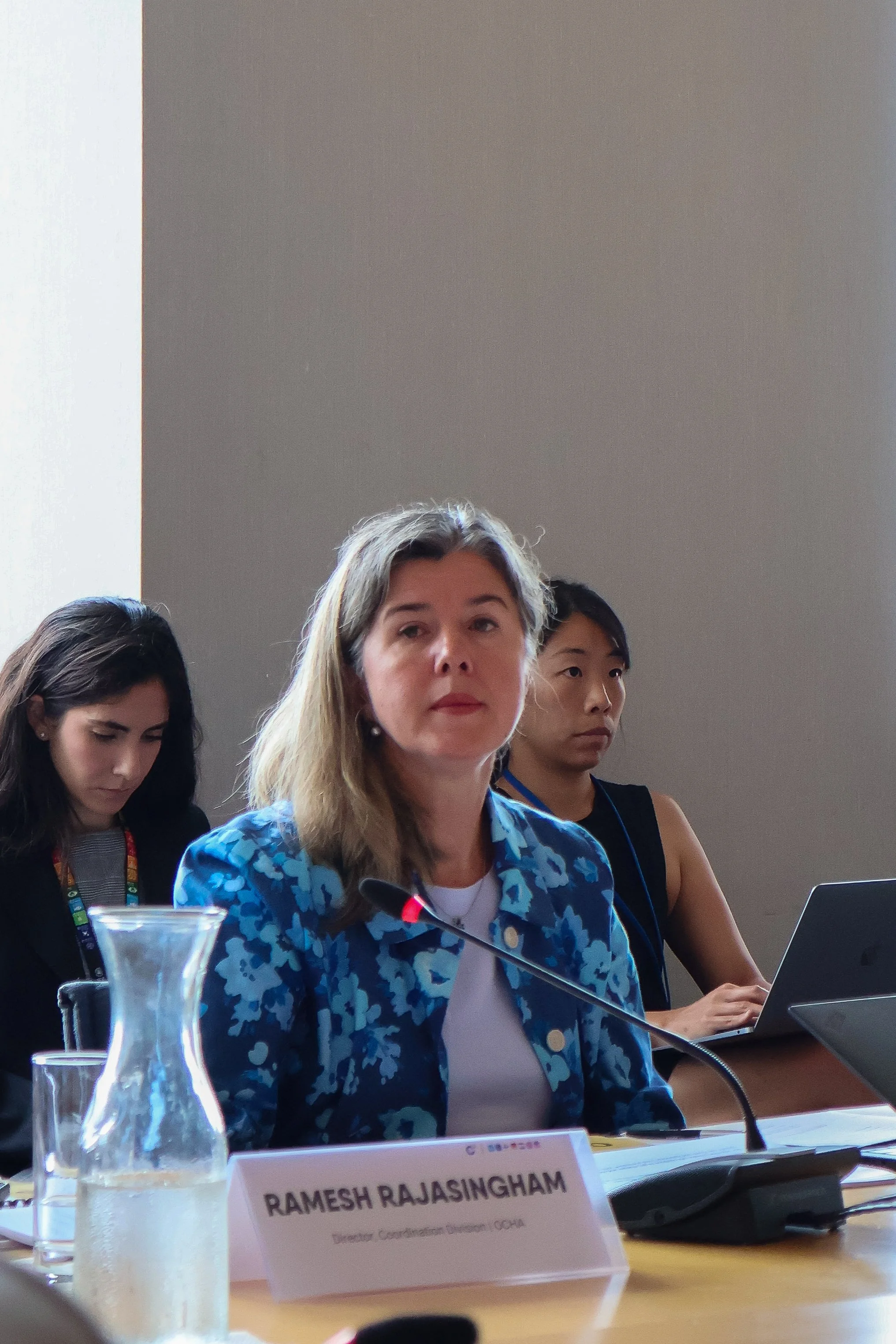 A woman with shoulder-length blonde hair wearing a blue patterned blazer sitting at a conference table with a nameplate that reads RAMESH RAJASINGHAM. She is speaking into a microphone. Two women are sitting behind her, one working on a laptop. There