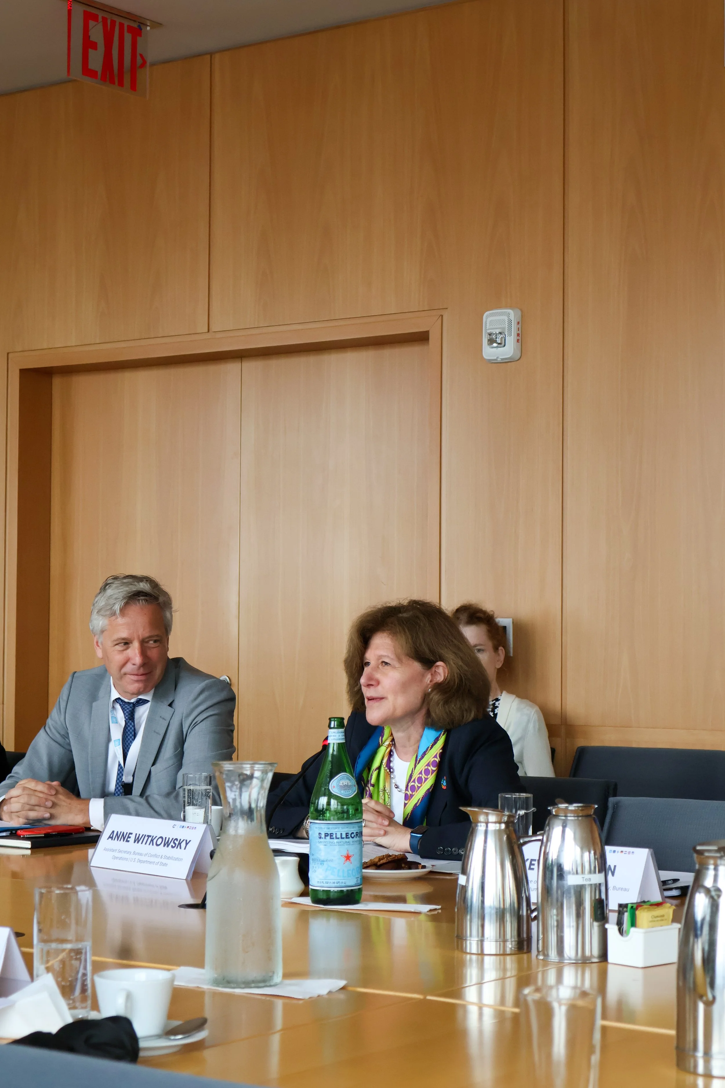 A woman speaking at a conference table with bottles of water, a pitcher, and coffee cups. She is wearing a dark blazer and a colorful scarf. Two other people are partially visible behind her, and an exit sign is above in the background.