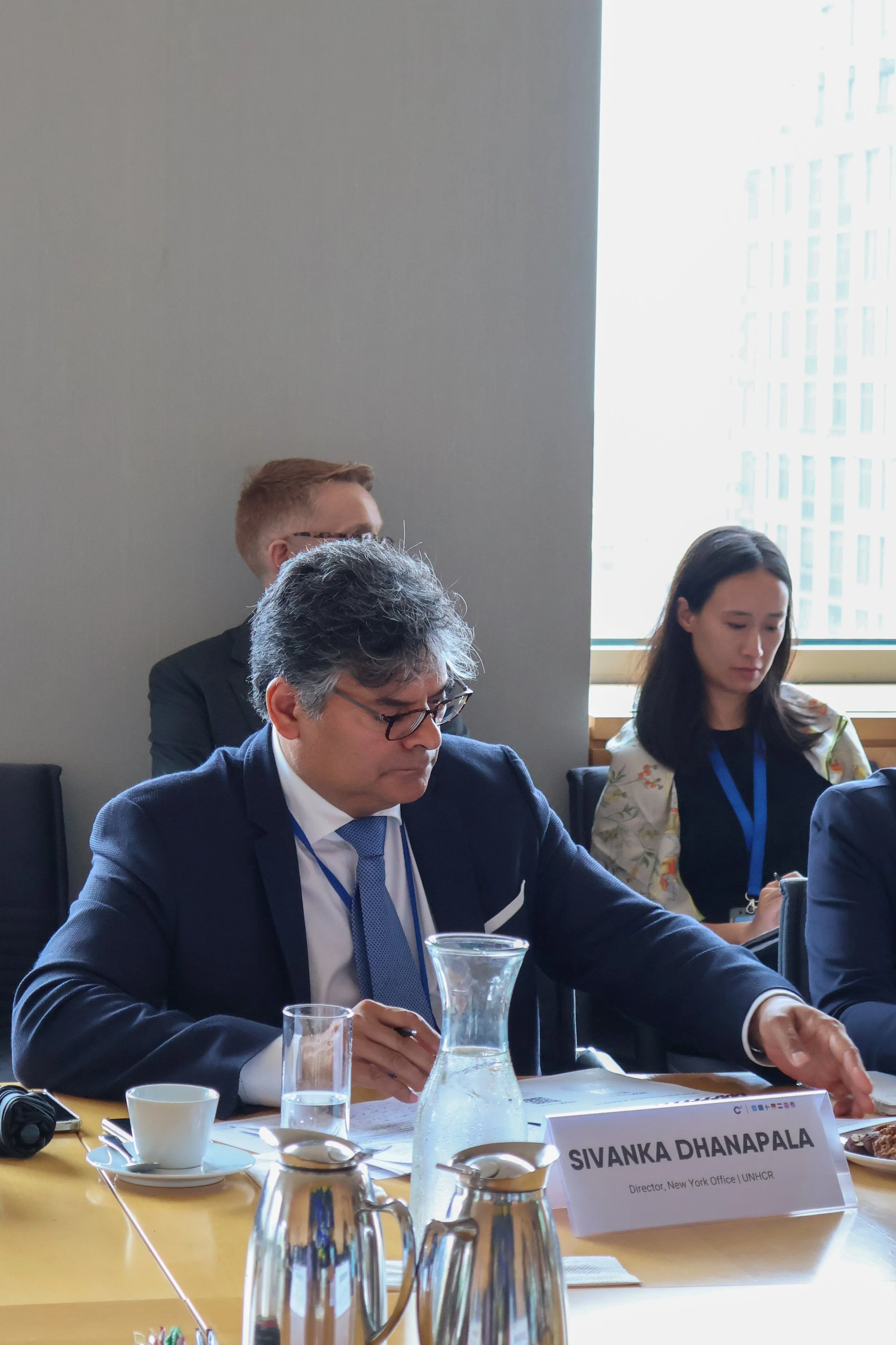 A group of people at a conference table with a nameplate that reads "SIVANKA DHANAPALA, Director, New York Office, UNHCR." The table has a water pitcher, glasses, and coffee cups. The individuals are engaged in a meeting or discussion in a bright roo