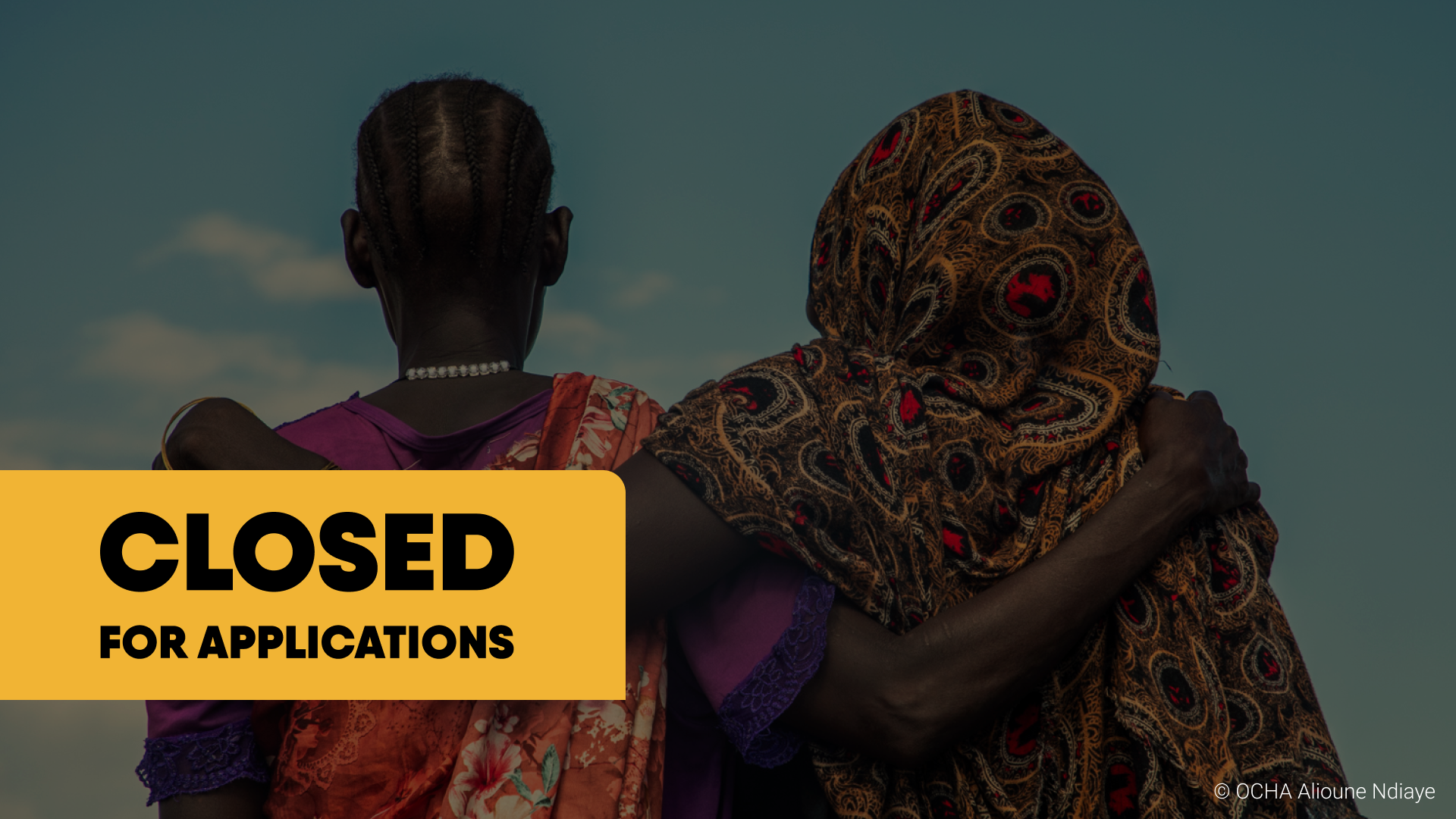 Two women embracing, one in a colorful headscarf and dress, the other in a purple dress, with a cloudy sky background. A yellow banner with black text over the image reads 'CLOSED FOR APPLICATIONS'.