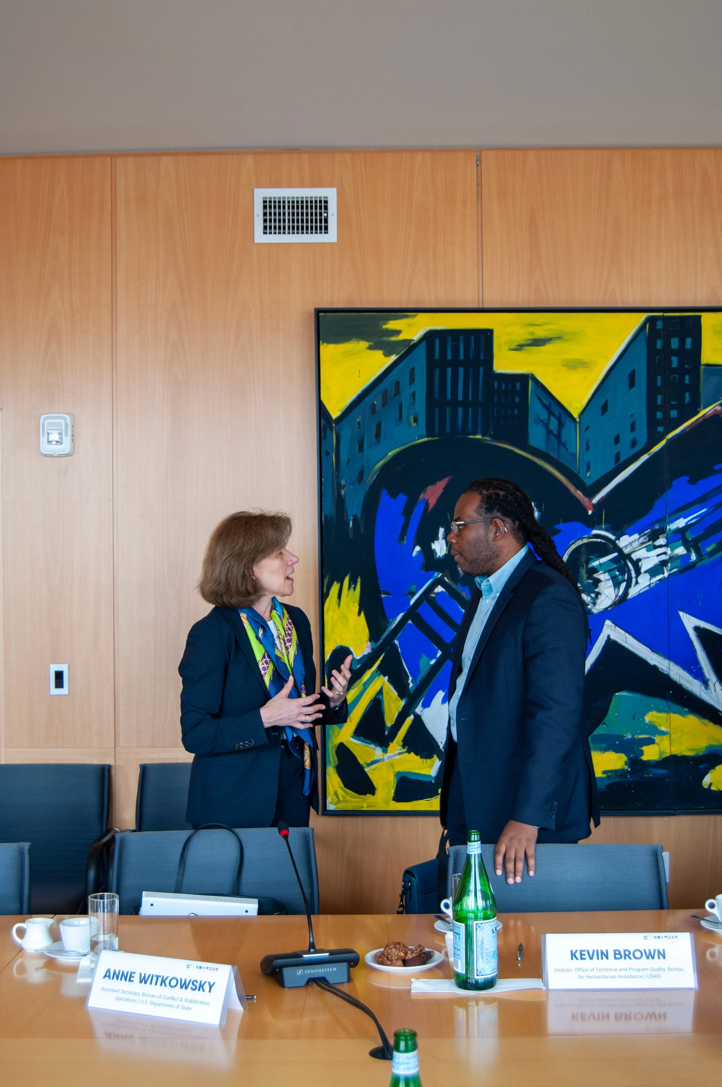 Two people, a woman and a man, are talking in a conference room. The woman is gesturing with her hands while the man listens. There are name tags on the table reading 'Anne Witkowsky' and 'Kevin Brown.' The background features a colorful abstract pai