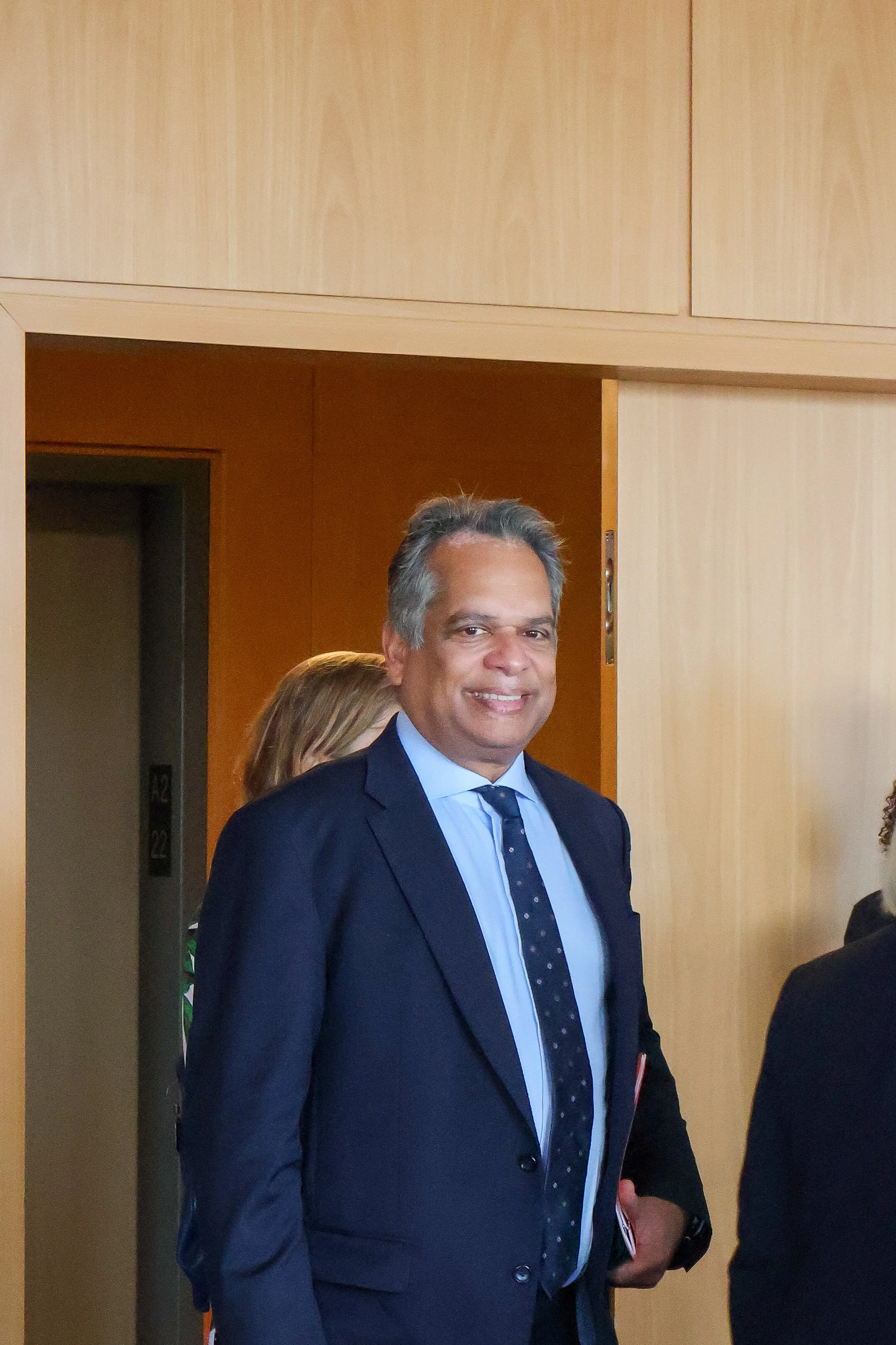 A smiling man in a dark suit and light blue shirt, standing indoors near an open doorway, with a woman partially visible behind him.