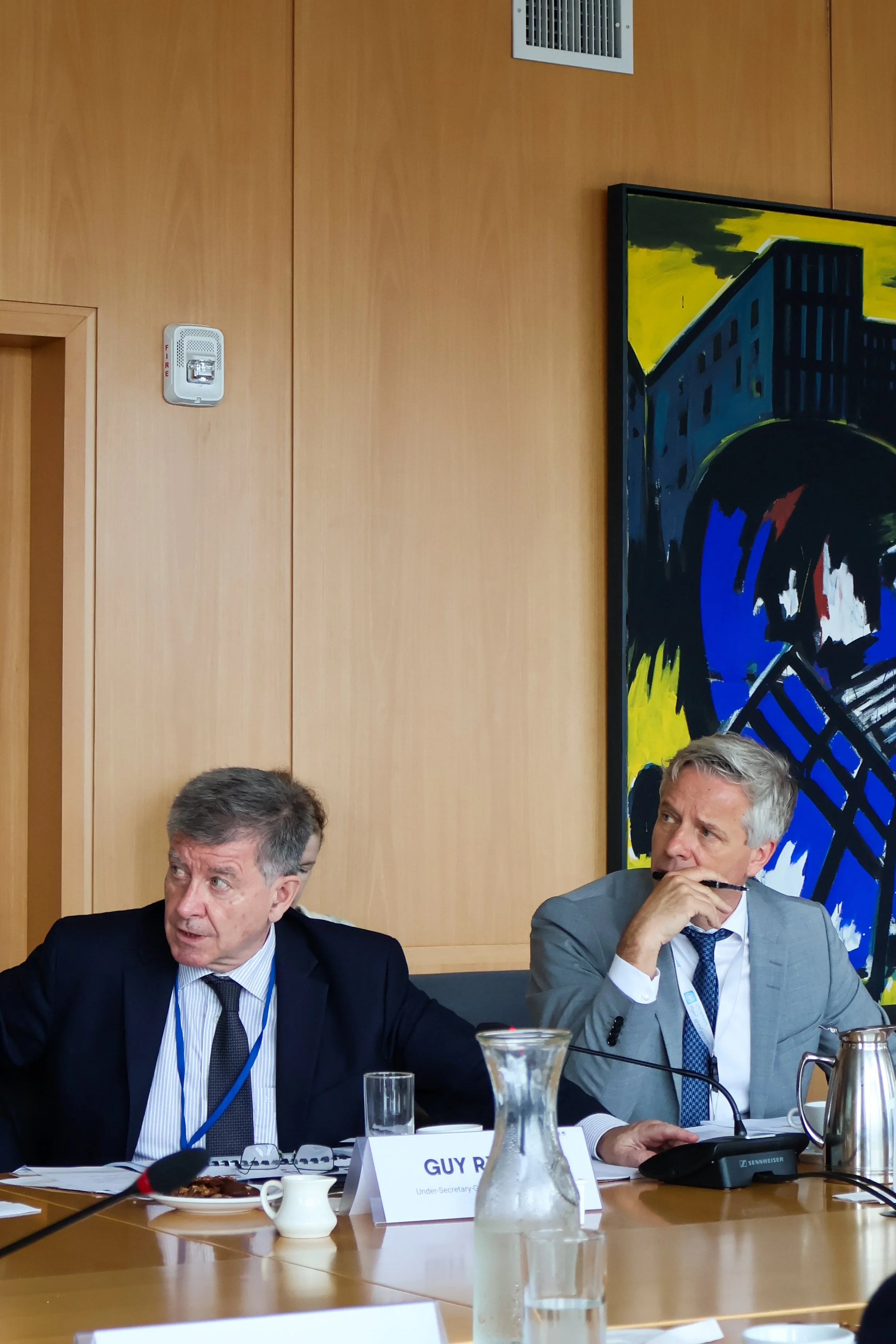 Two men in suits seated at a conference table during a meeting, with nameplates, microphones, a pitcher of water, and a painting on the wall behind them.