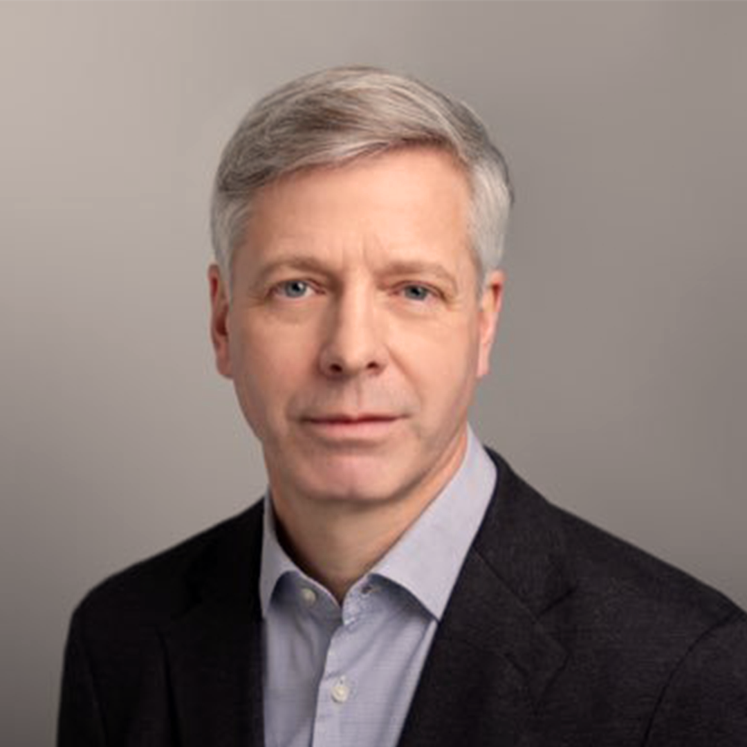 Headshot of a middle-aged man with gray hair, wearing a dark suit jacket and light-colored dress shirt, against a neutral background.