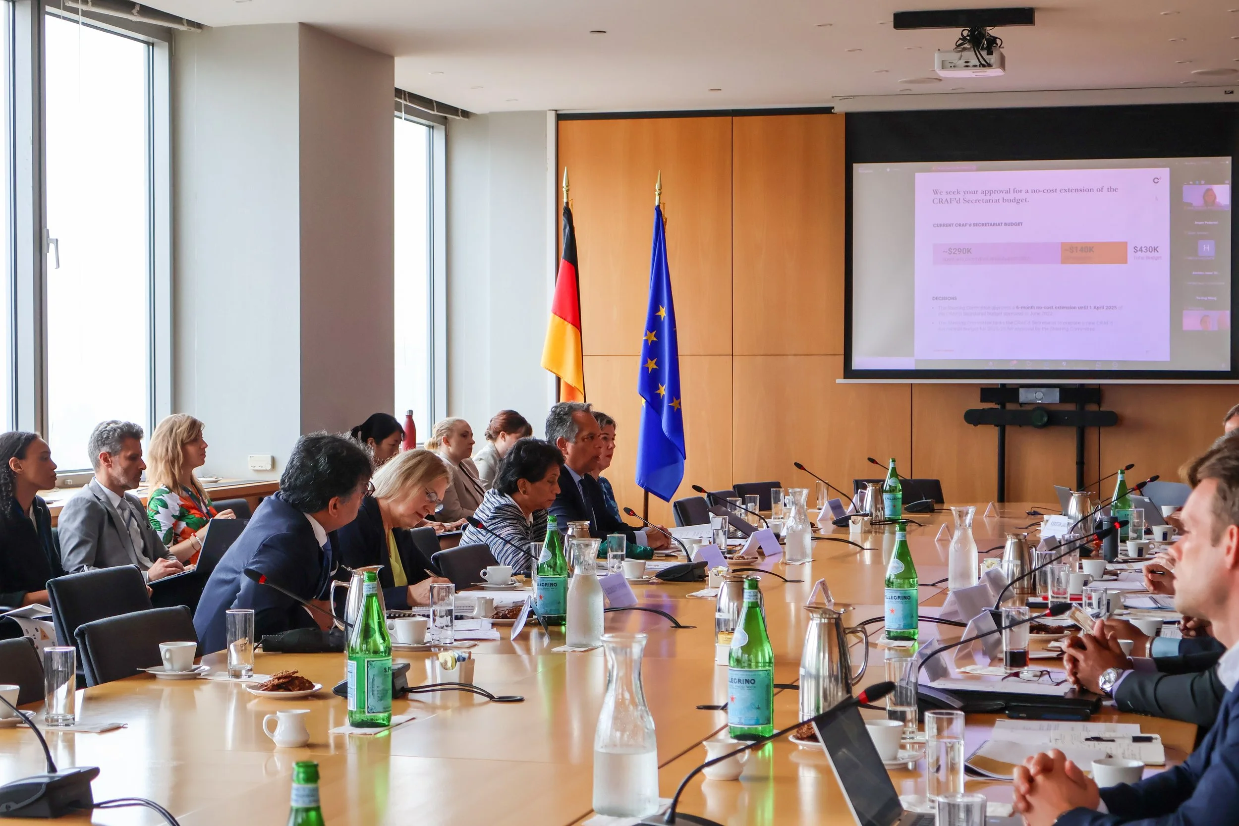 A conference room with several people seated at a large table during a meeting. The room has large windows, and flags of Germany and the European Union are displayed in front of a wooden wall. A large screen shows a presentation related to a budget e